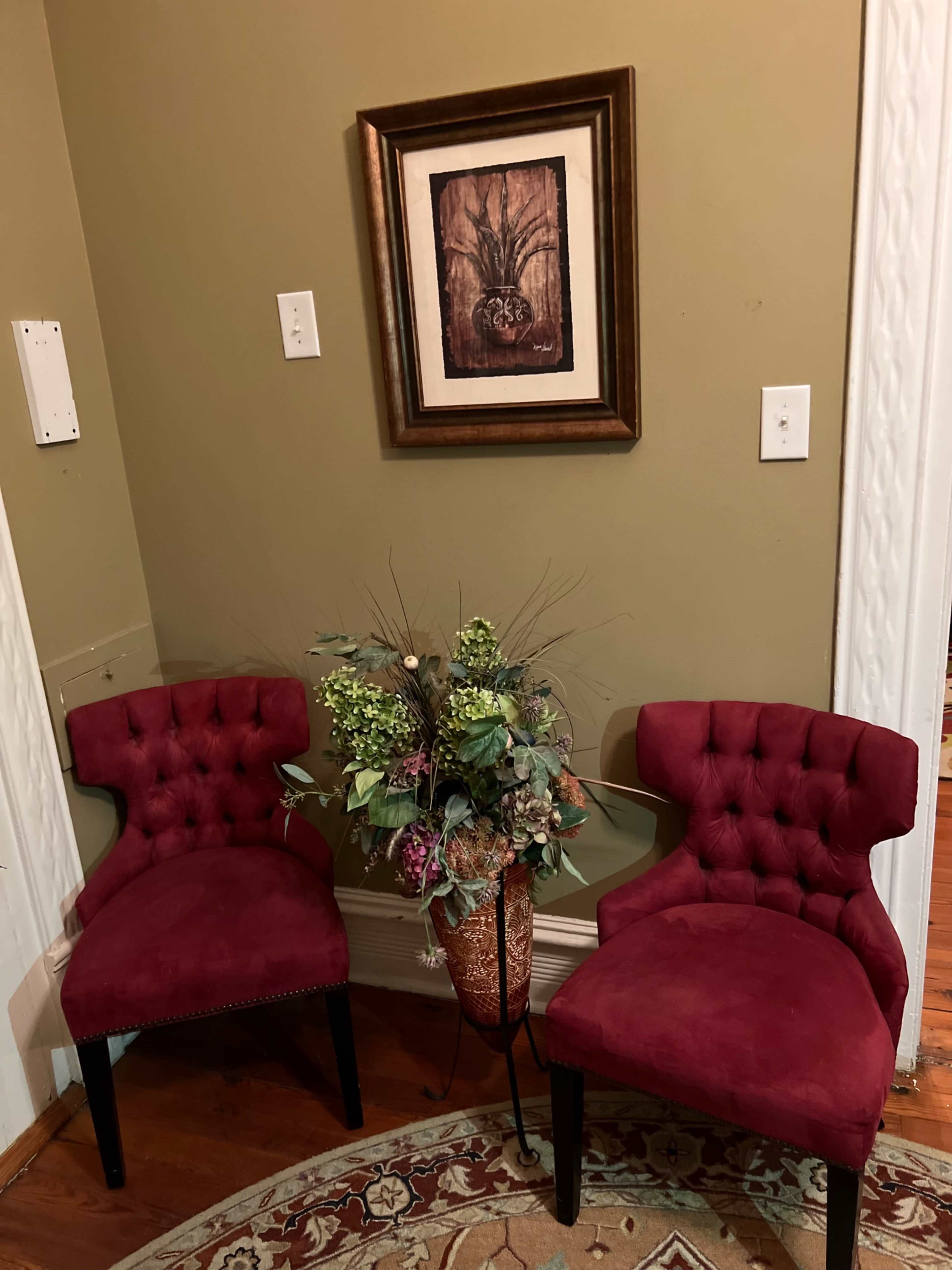 A small corner with two red tufted chairs flanking a floral arrangement in a metallic vase, set against a green wall and a framed artwork above.