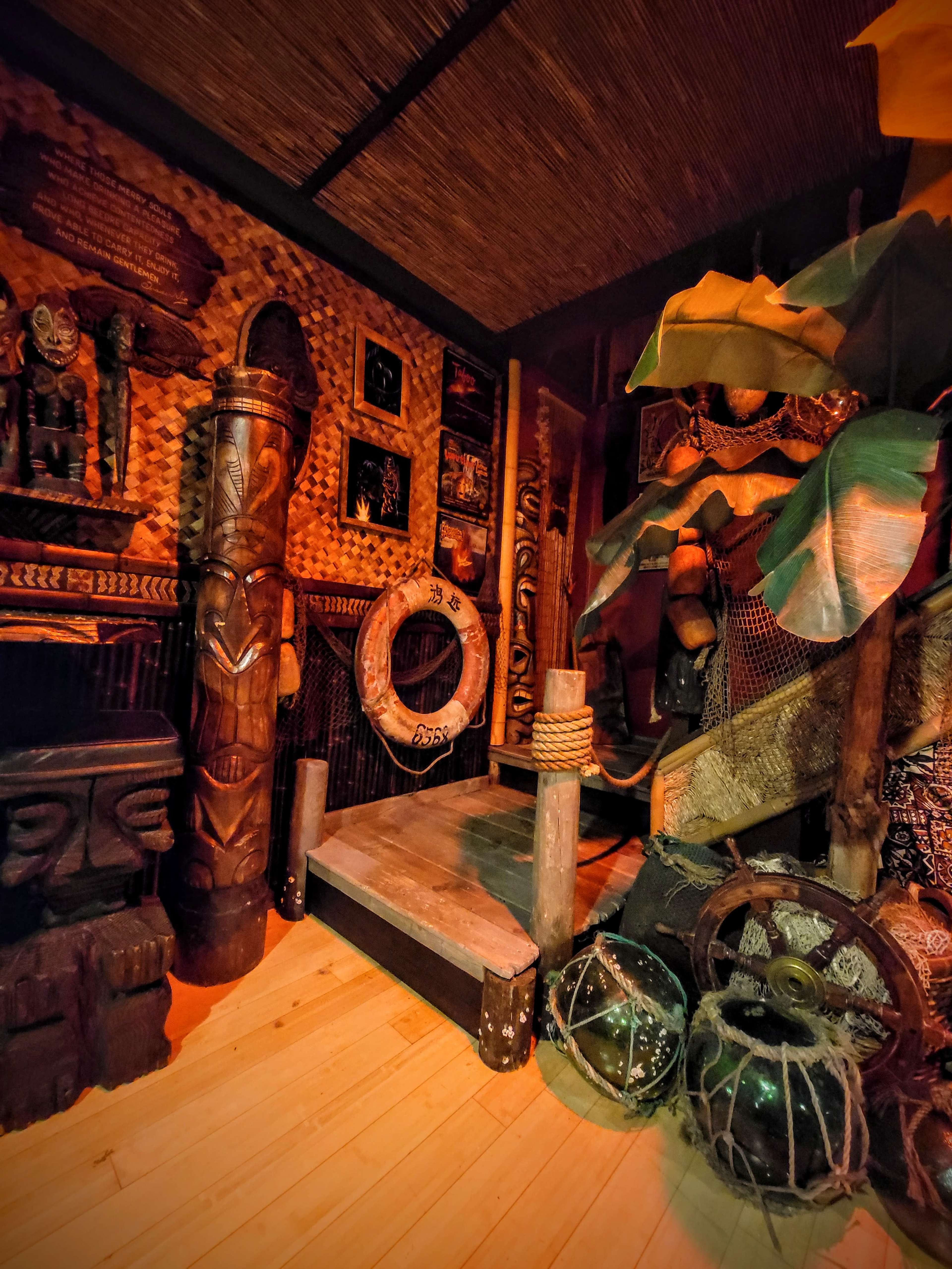The image depicts a dimly lit room featuring a wooden platform surrounded by tropical decorations, carved wooden figures, and various artifacts.