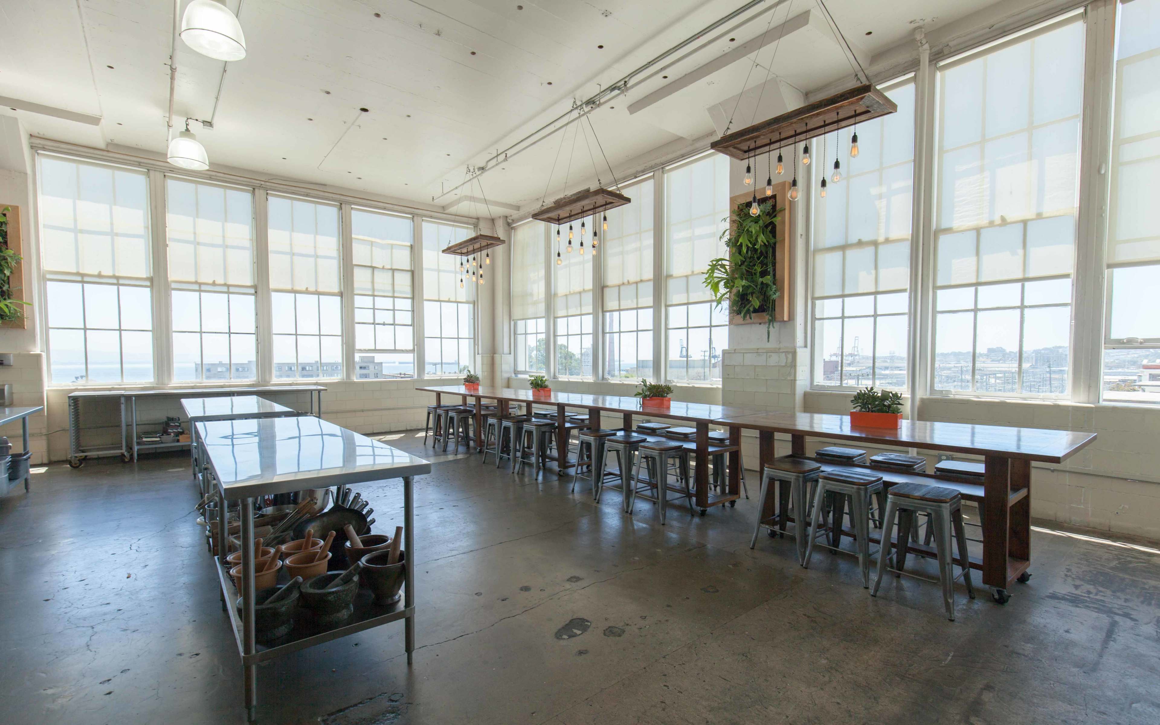 A spacious kitchen with large windows, long wooden tables, metal stools, and ceiling-mounted plants, all set against a concrete floor.