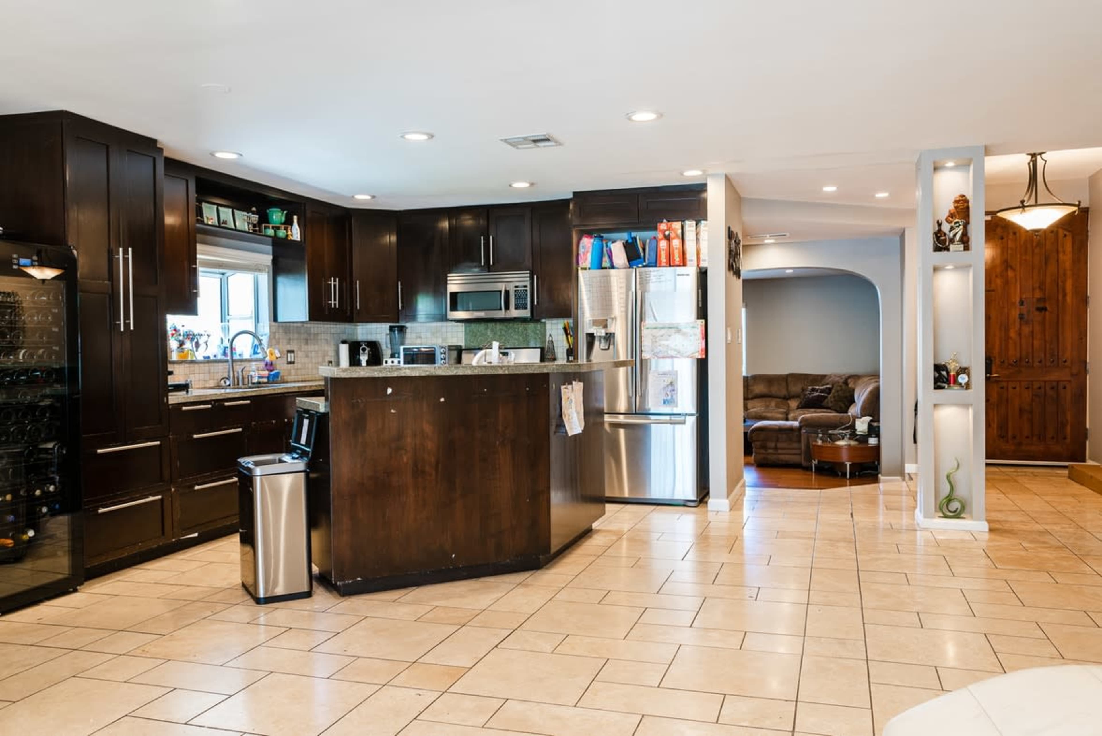 The image shows a modern kitchen with dark wood cabinets, stainless steel appliances, and a tile floor, opening into a living area.