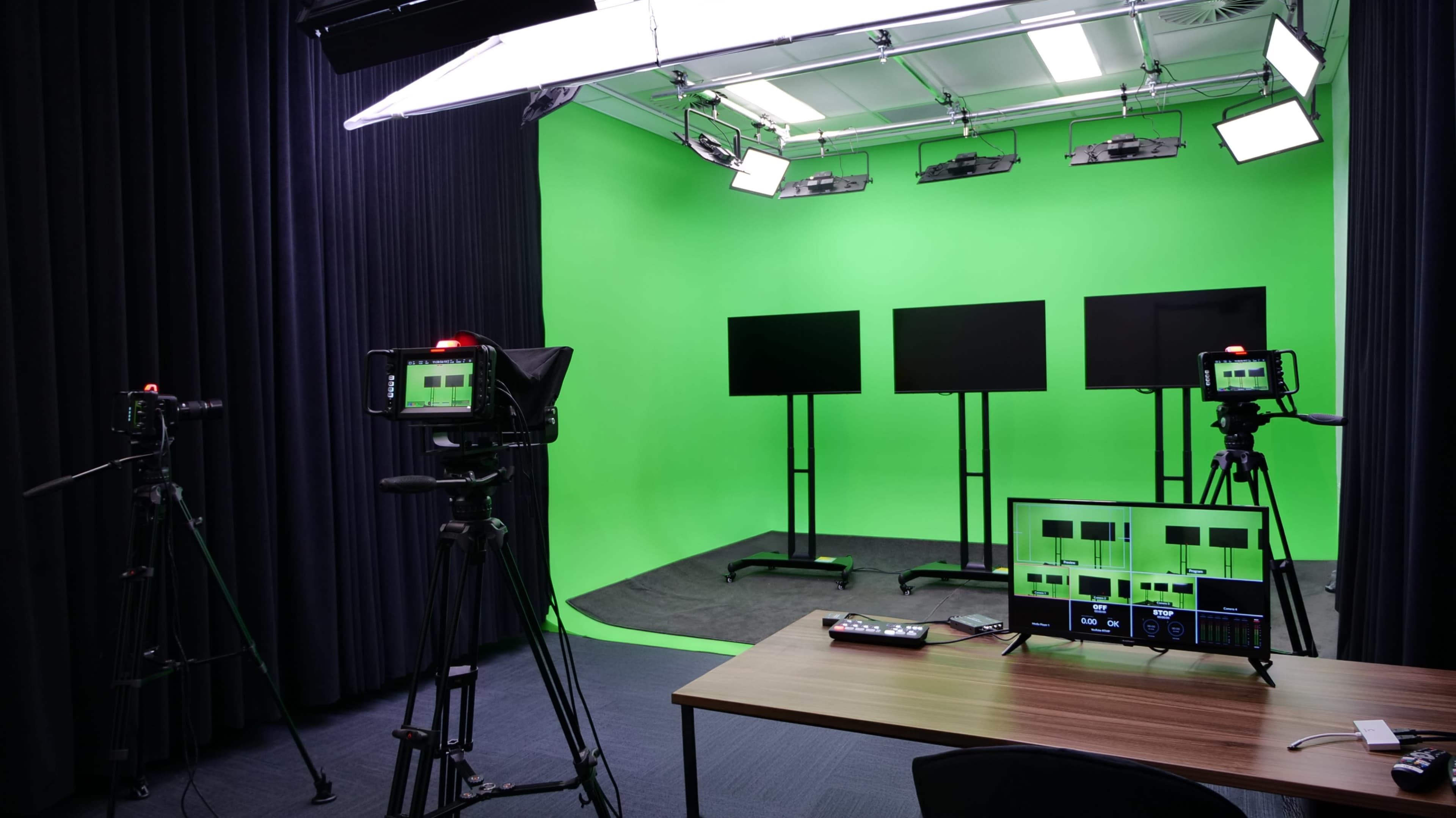 The image shows a studio setup with three black screens on stands, two video cameras, and a control desk, all against a green backdrop.