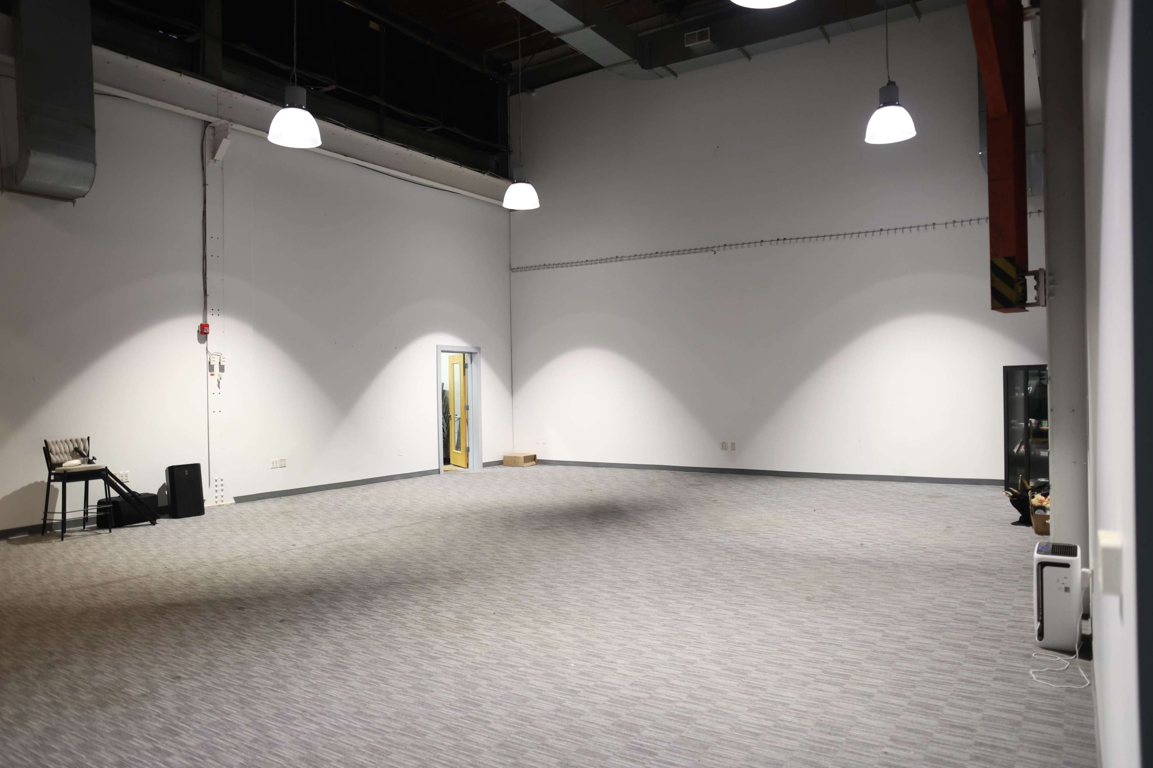 The image shows an empty, well-lit room with gray carpet and white walls, featuring a single door and a chair against one wall.