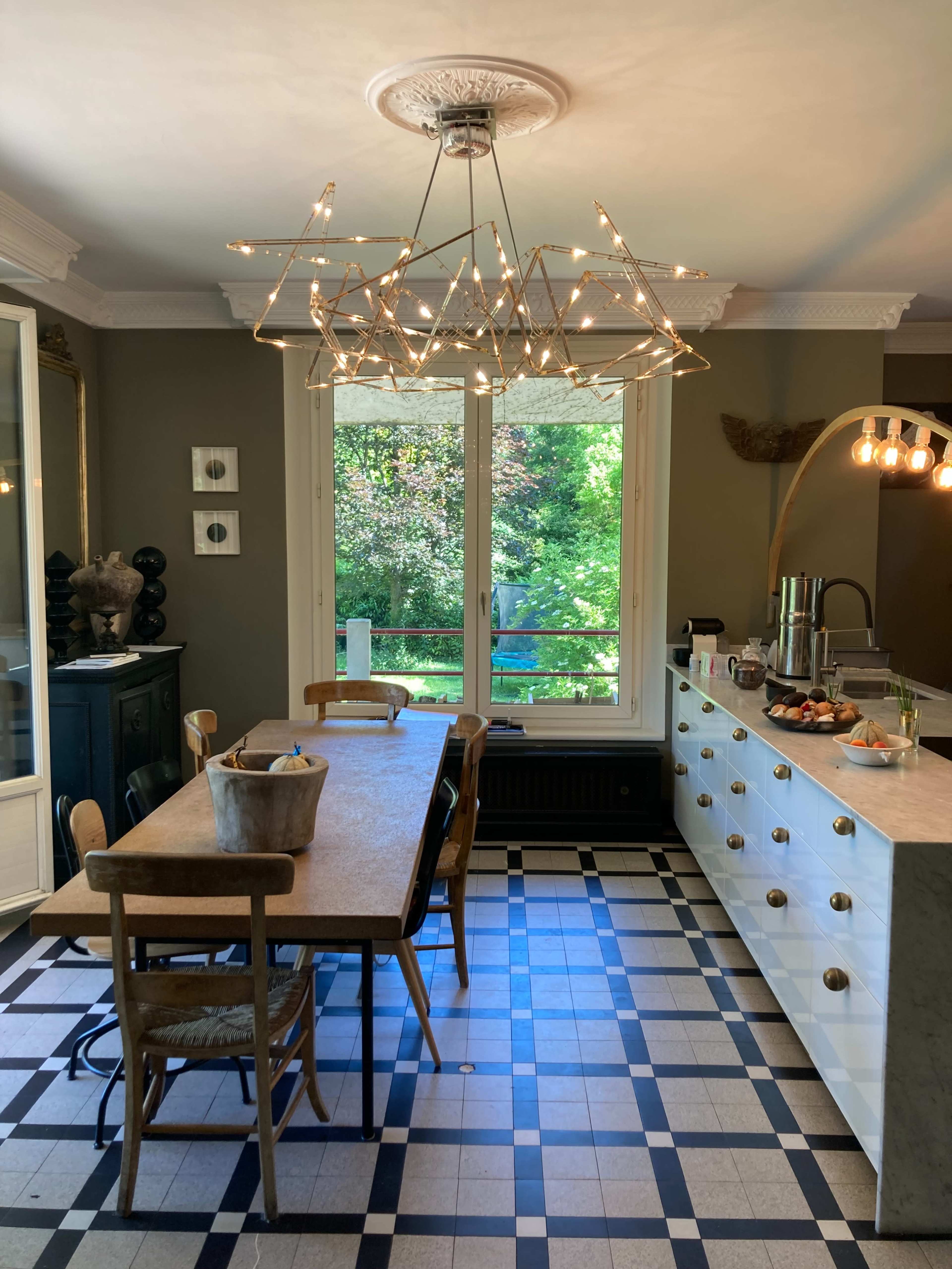 The kitchen features a large wooden dining table, a modern chandelier, and a view of a green garden through a set of windows.