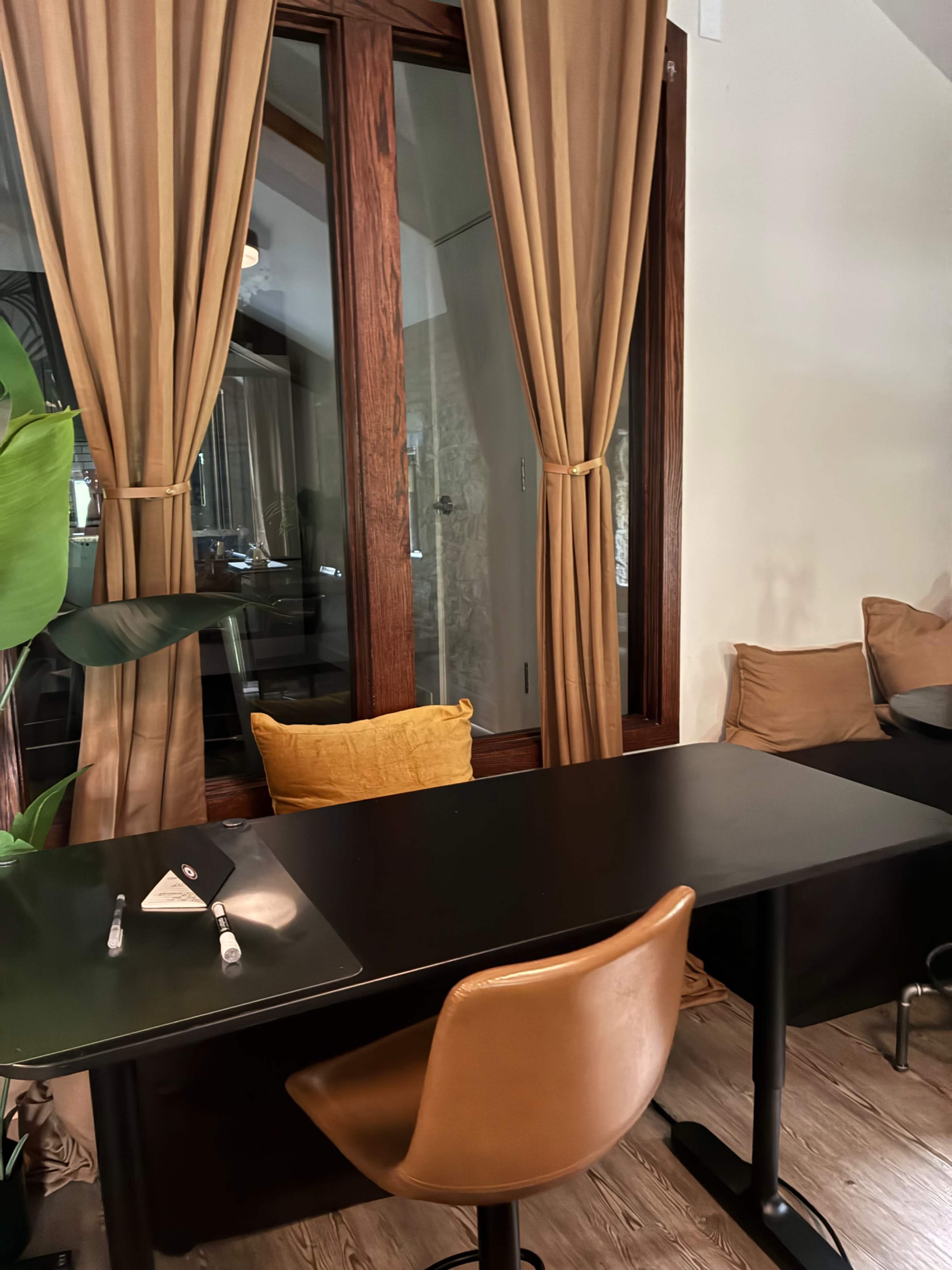 A simple black desk with a brown chair sits beside a window adorned with beige curtains, surrounded by cushions and indoor plants.