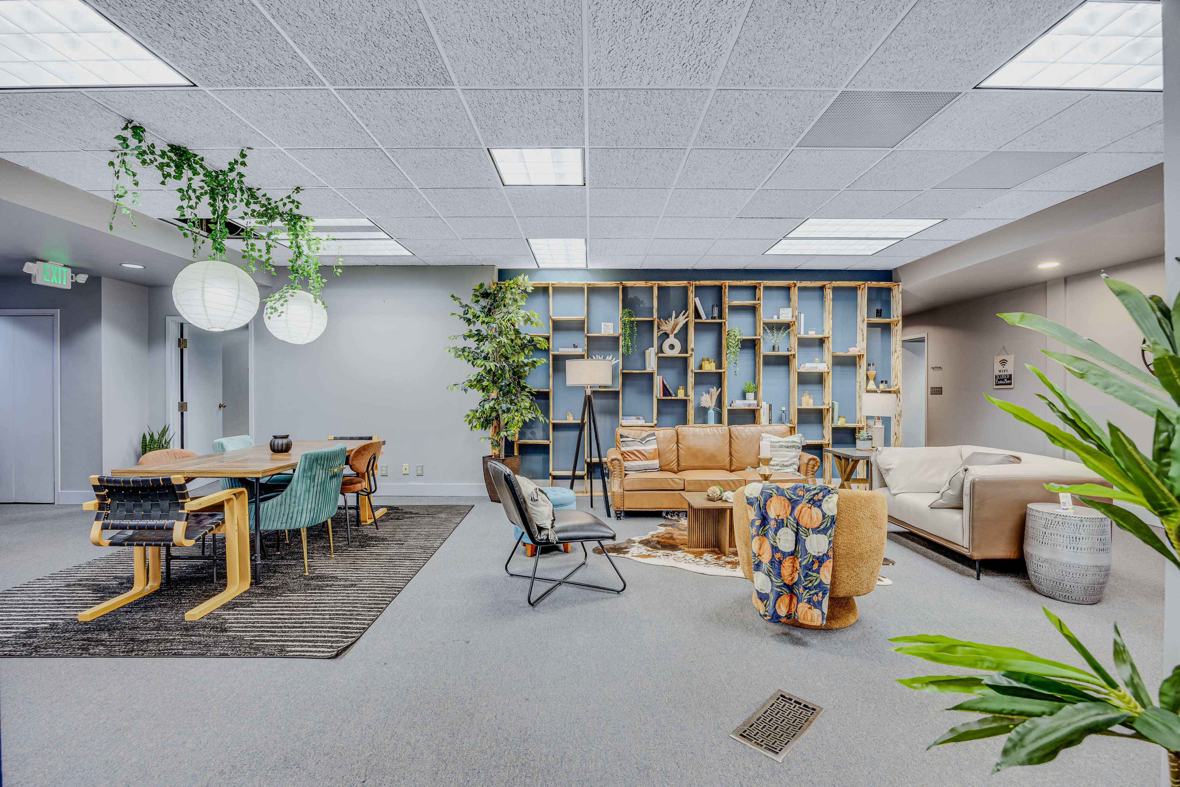 The image shows a modern open workspace featuring a mix of seating areas, a dining table, plants, and decorative bookshelves.