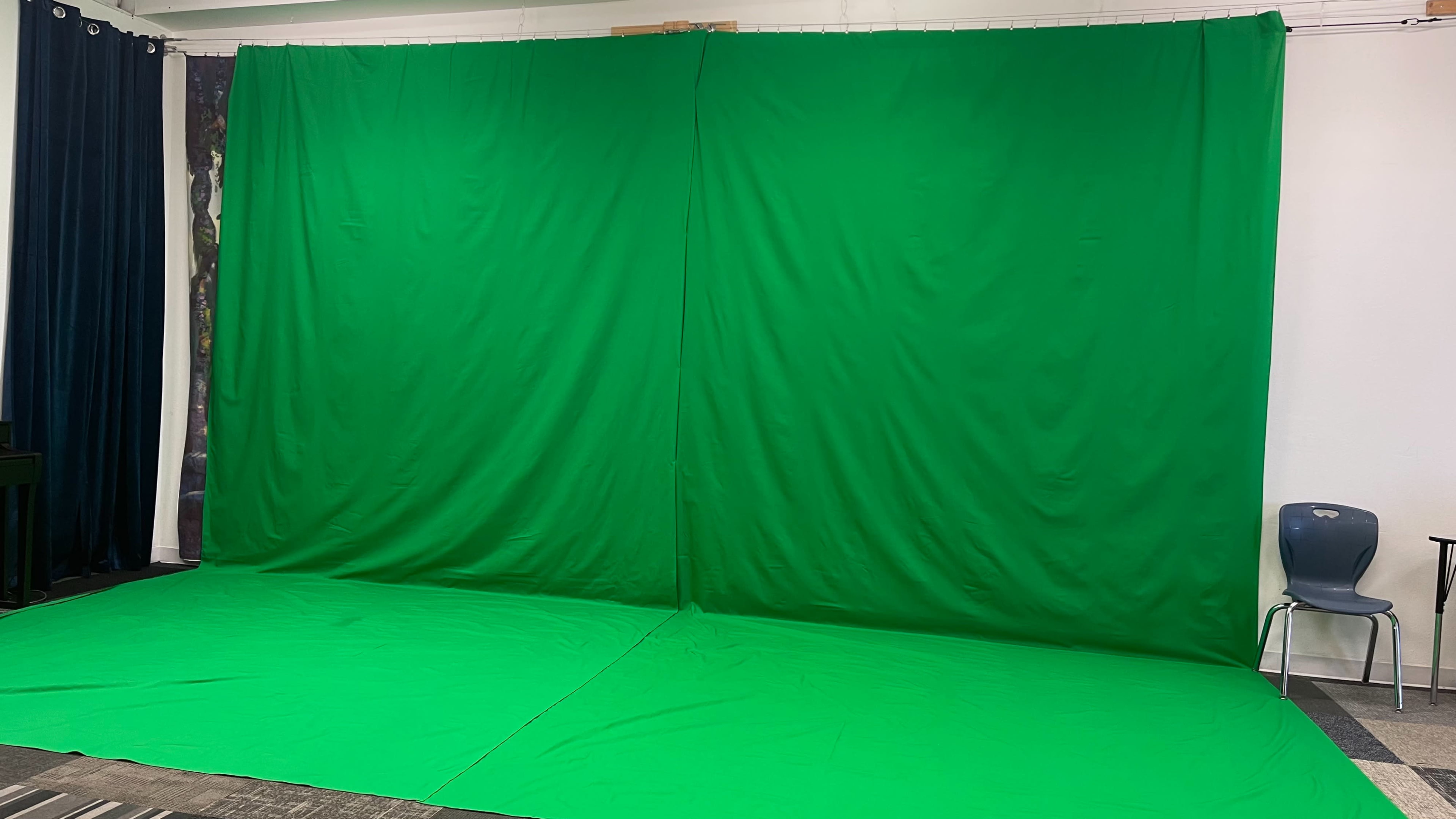 A large green screen set up against a wall with a chair and a table visible in the background.