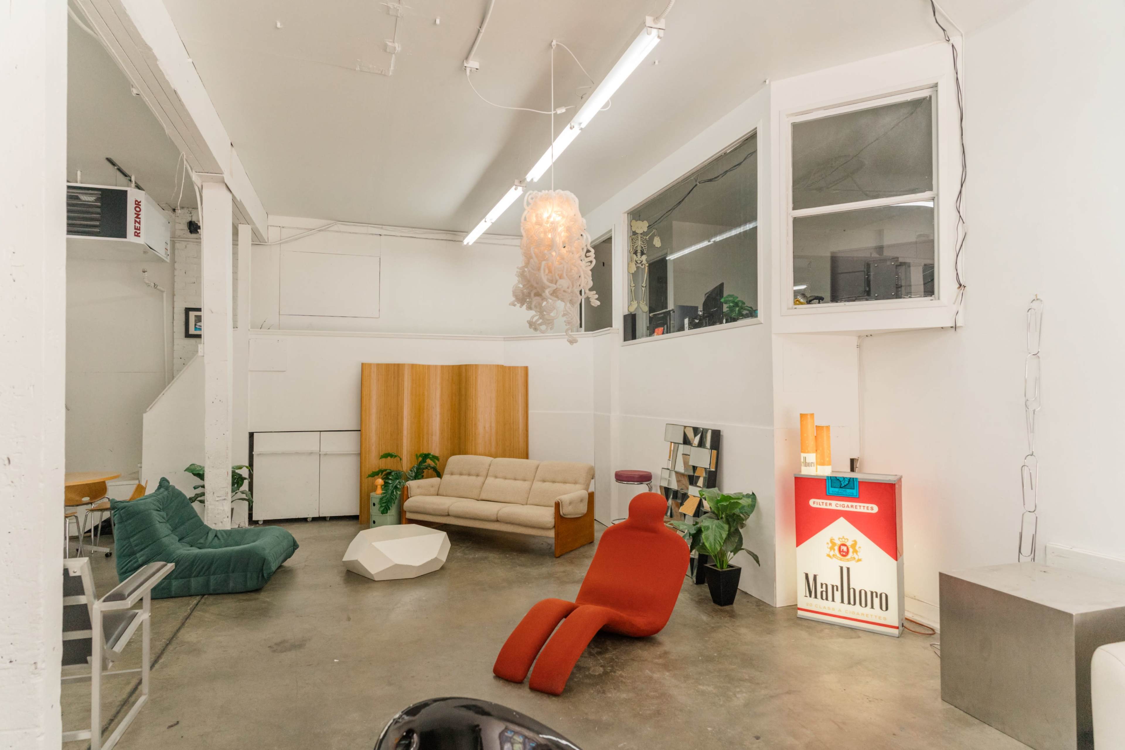 An interior space features a mix of modern furniture, including a beige sofa, green and red chairs, potted plants, and a Marlboro-branded display.