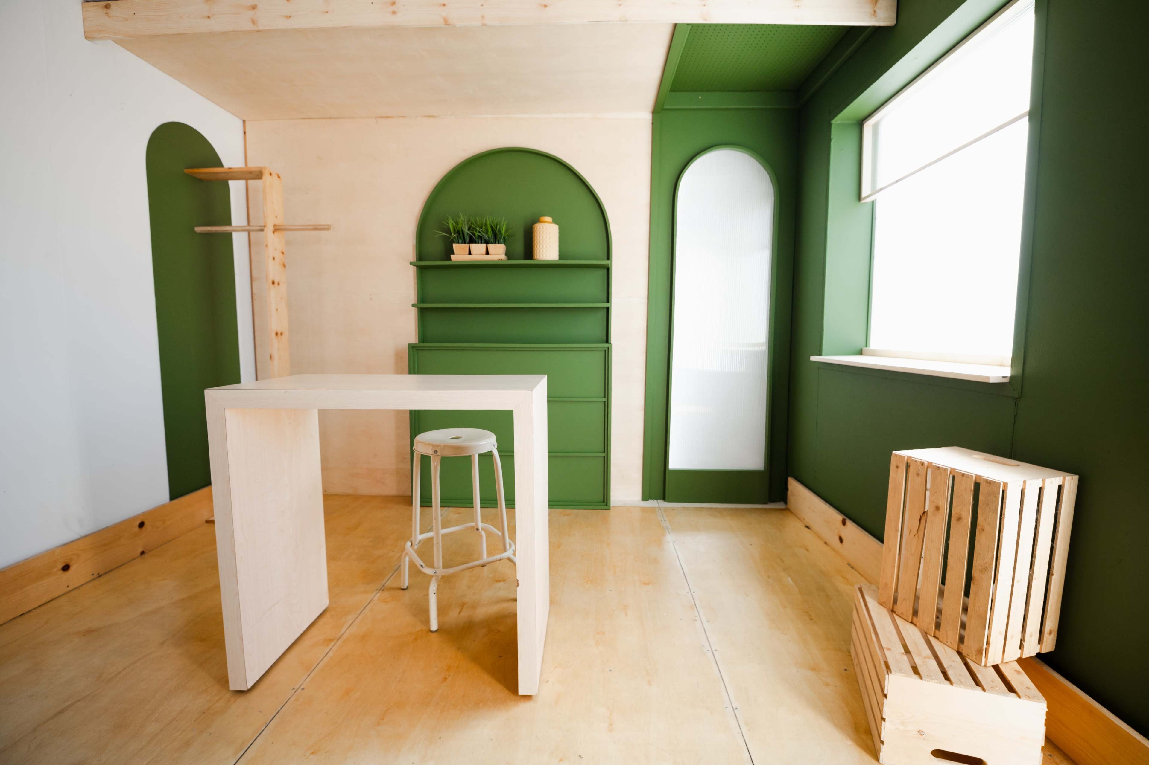 A minimalist room with a light wood floor, a white table and stool, green accent walls, and shelves with plants.