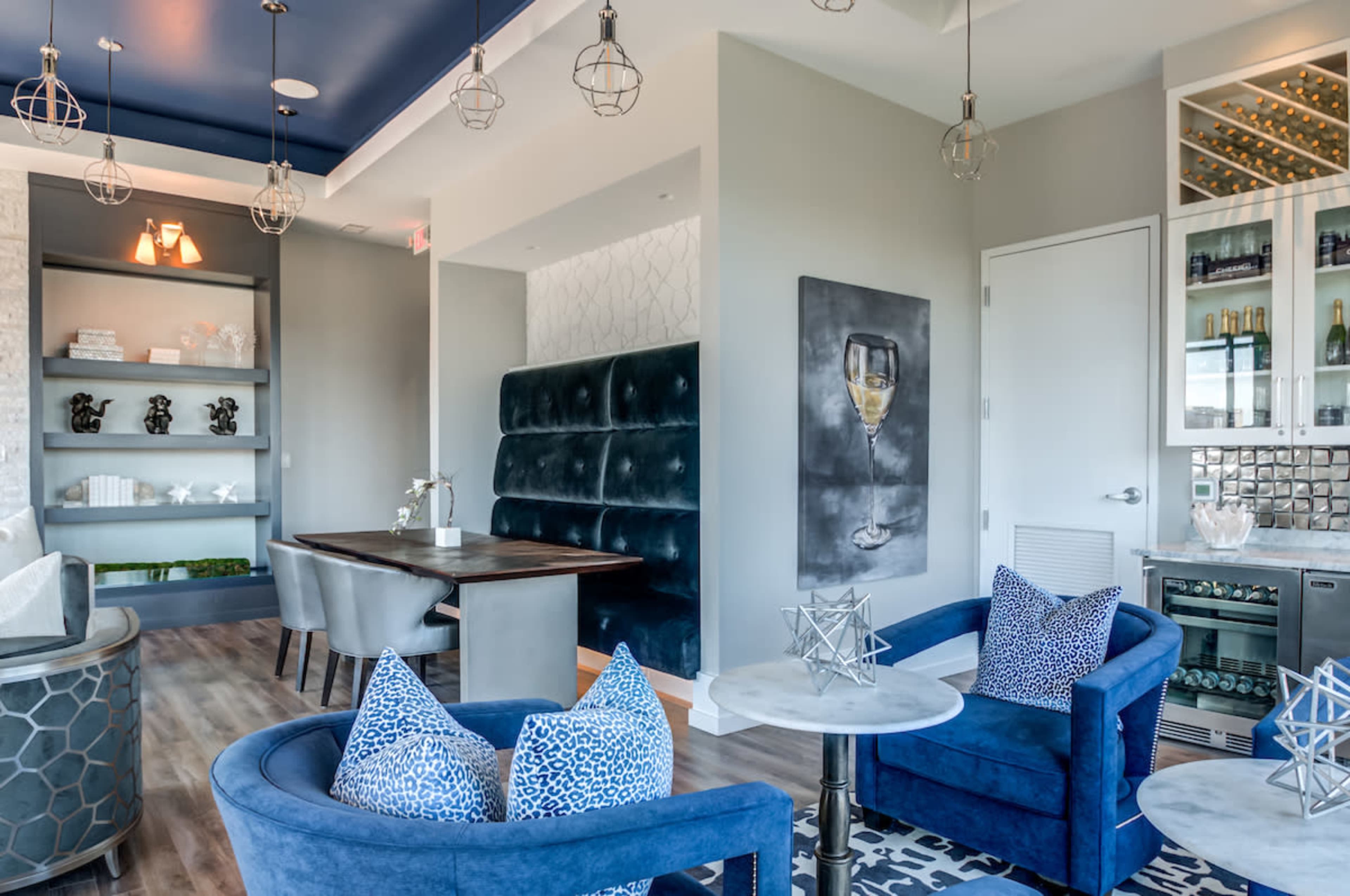 The image shows a modern lounge area featuring blue velvet chairs, a wooden table, and a bar cabinet with glass shelves amid contemporary decor.