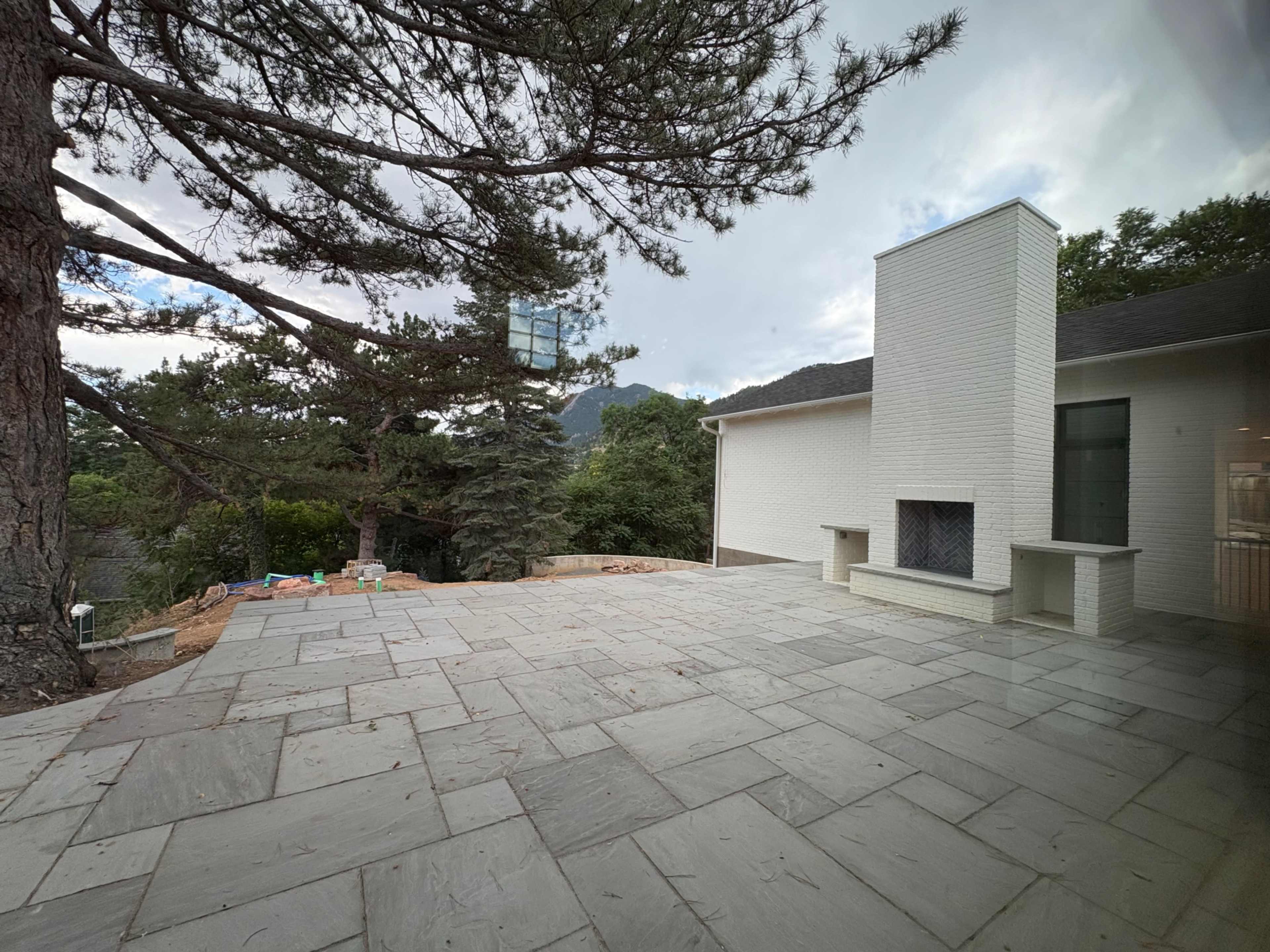 The image shows a large stone patio in a residential outdoor area, featuring a white brick building and a chimney surrounded by trees and mountains in the background.