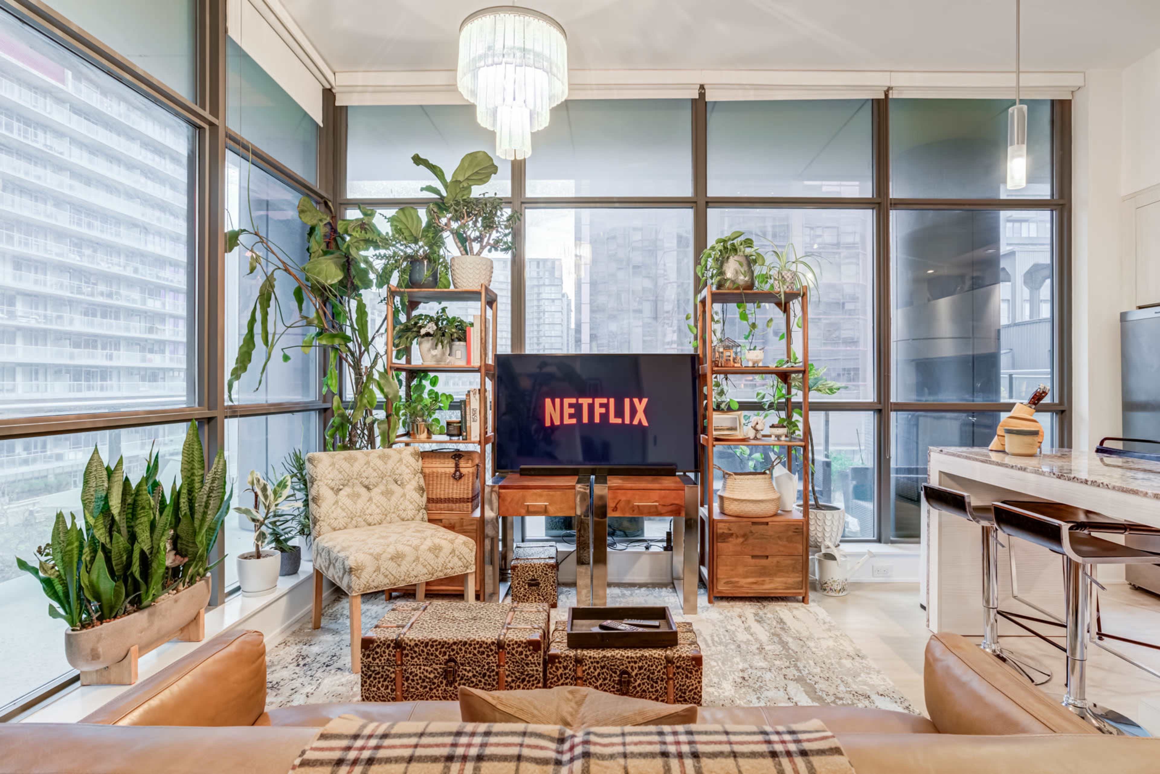 The living room features a large window with city views, a television displaying the Netflix logo, and plants integrated into wooden shelving, along with stylish seating and decor.