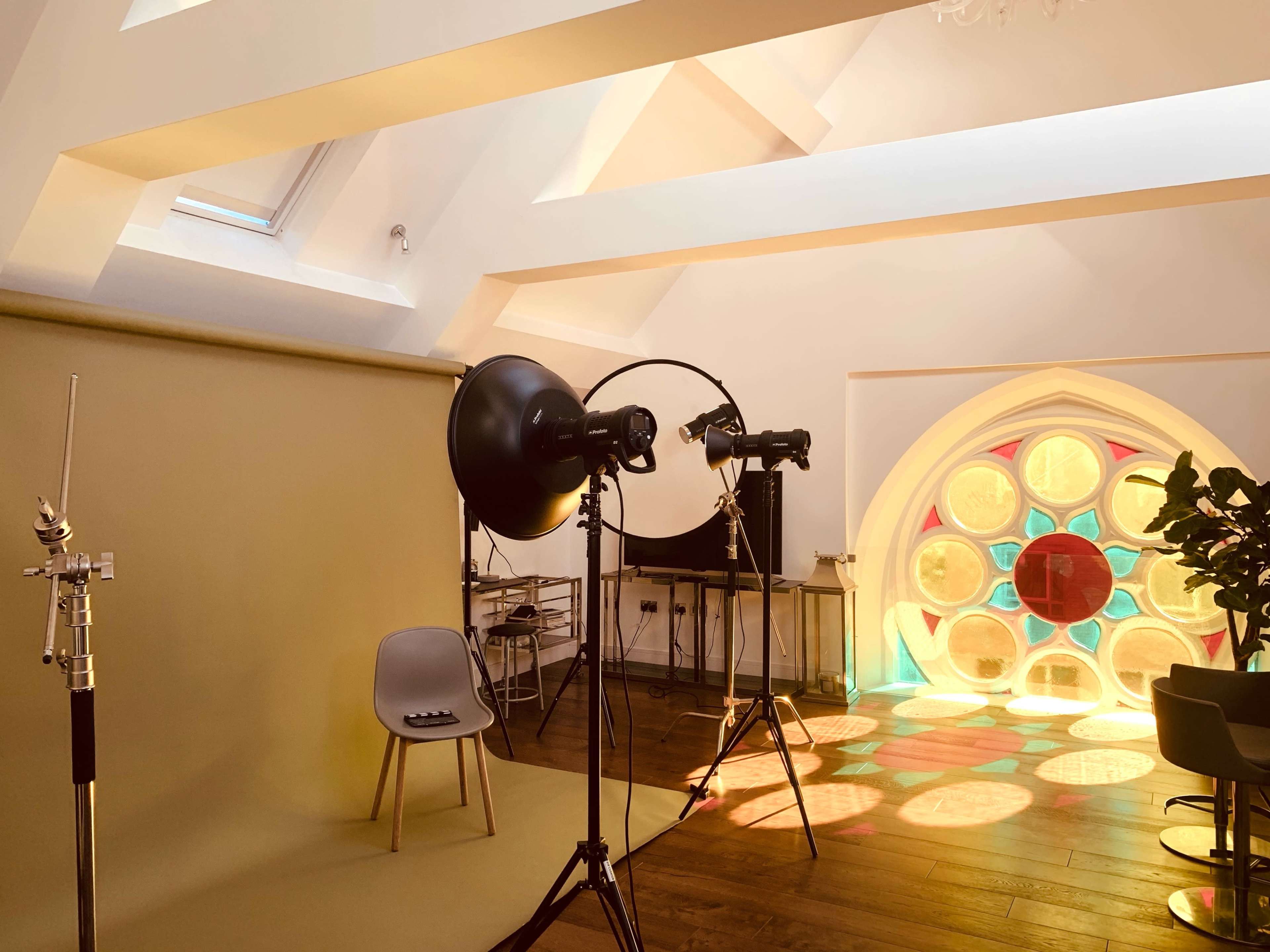 A photography studio is set up with a beige backdrop, lighting equipment, a chair, and a colorful window casting patterns on the floor.
