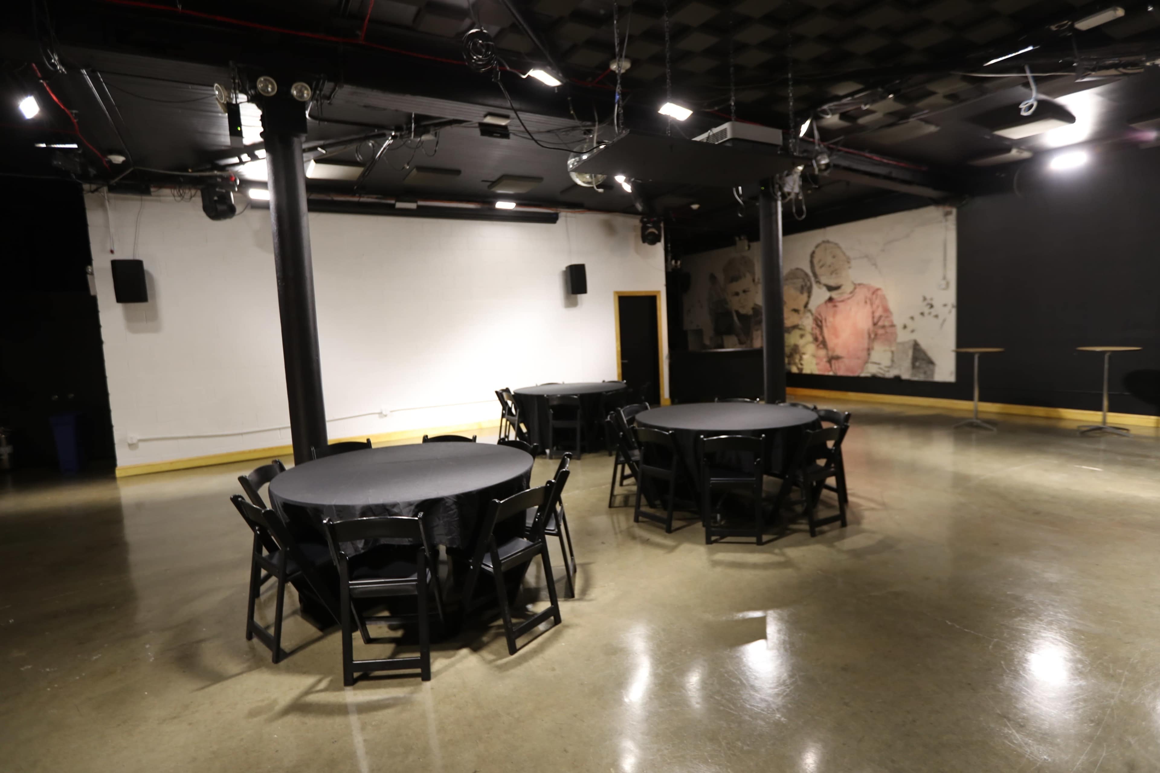 The image shows a venue with several round tables covered in black cloth, surrounded by empty space and illuminated by overhead lights.