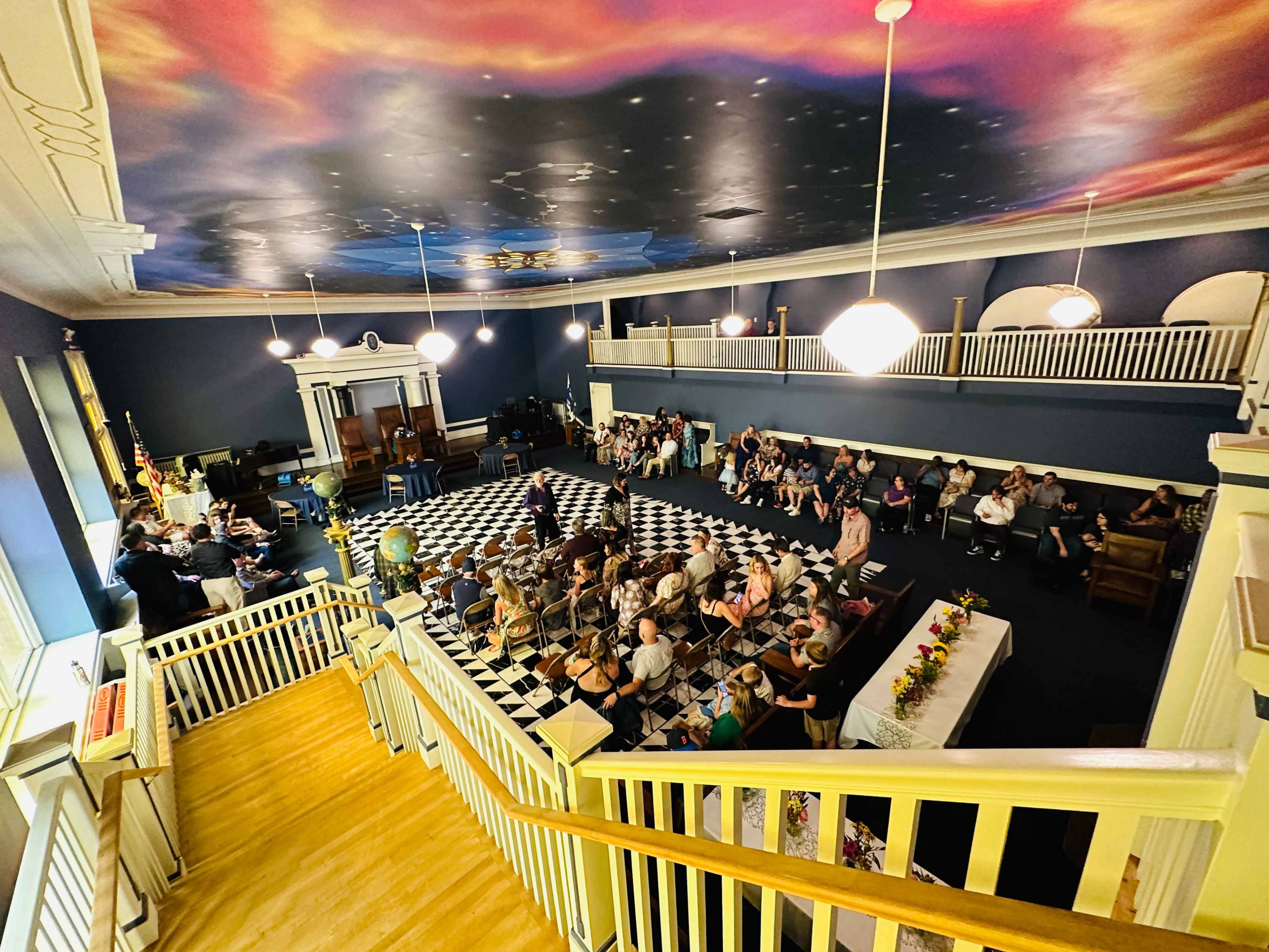 The image shows a large room with a checkered floor, filled with people seated in rows, and a stage area that includes wooden furniture and a colorful ceiling mural.