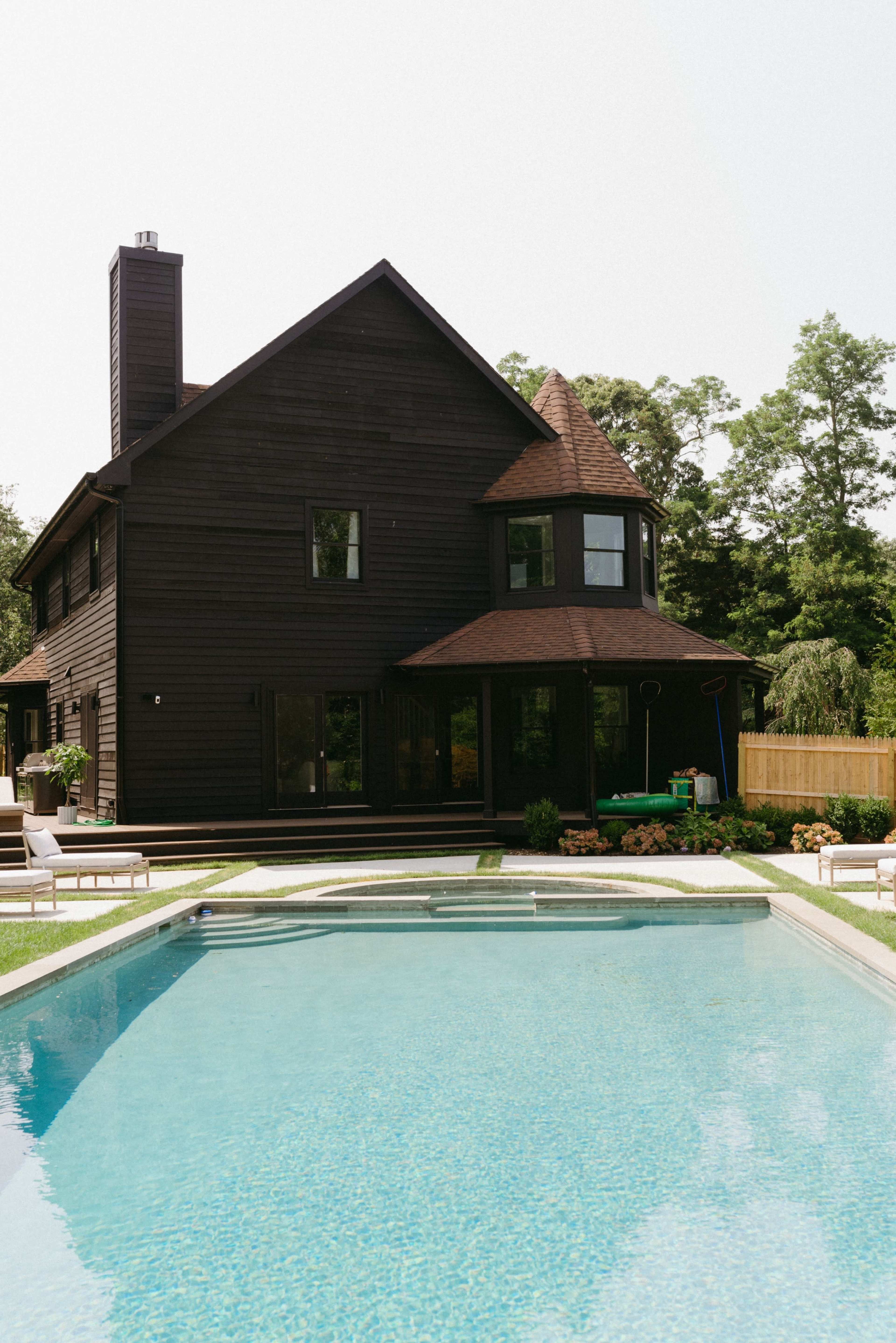 A modern, dark-colored house with multiple gables overlooks a swimming pool surrounded by a grassy area and lounge chairs.