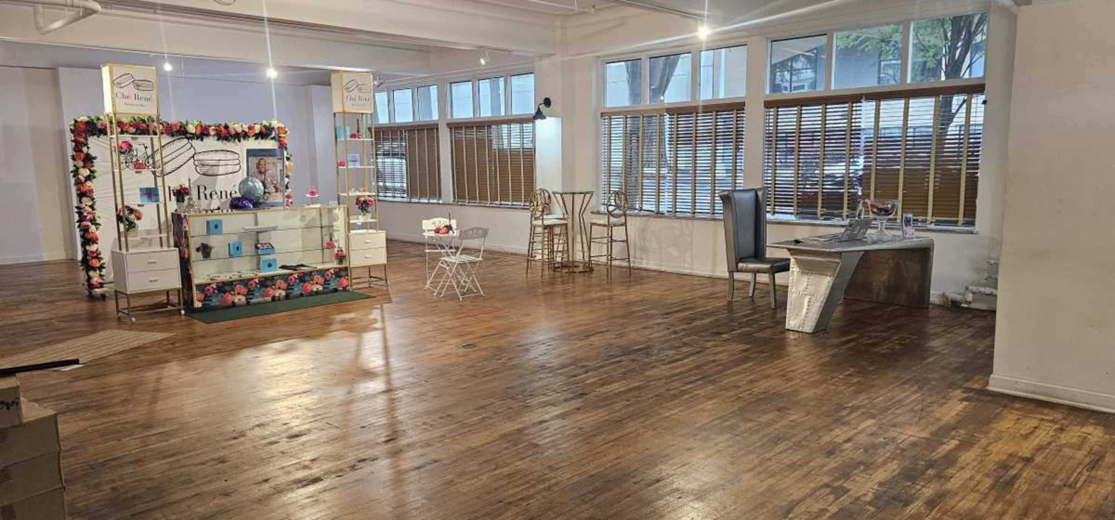 The image shows a spacious, partially decorated room with wooden floors, featuring a decorative display setup on one side and empty furniture pieces scattered throughout.