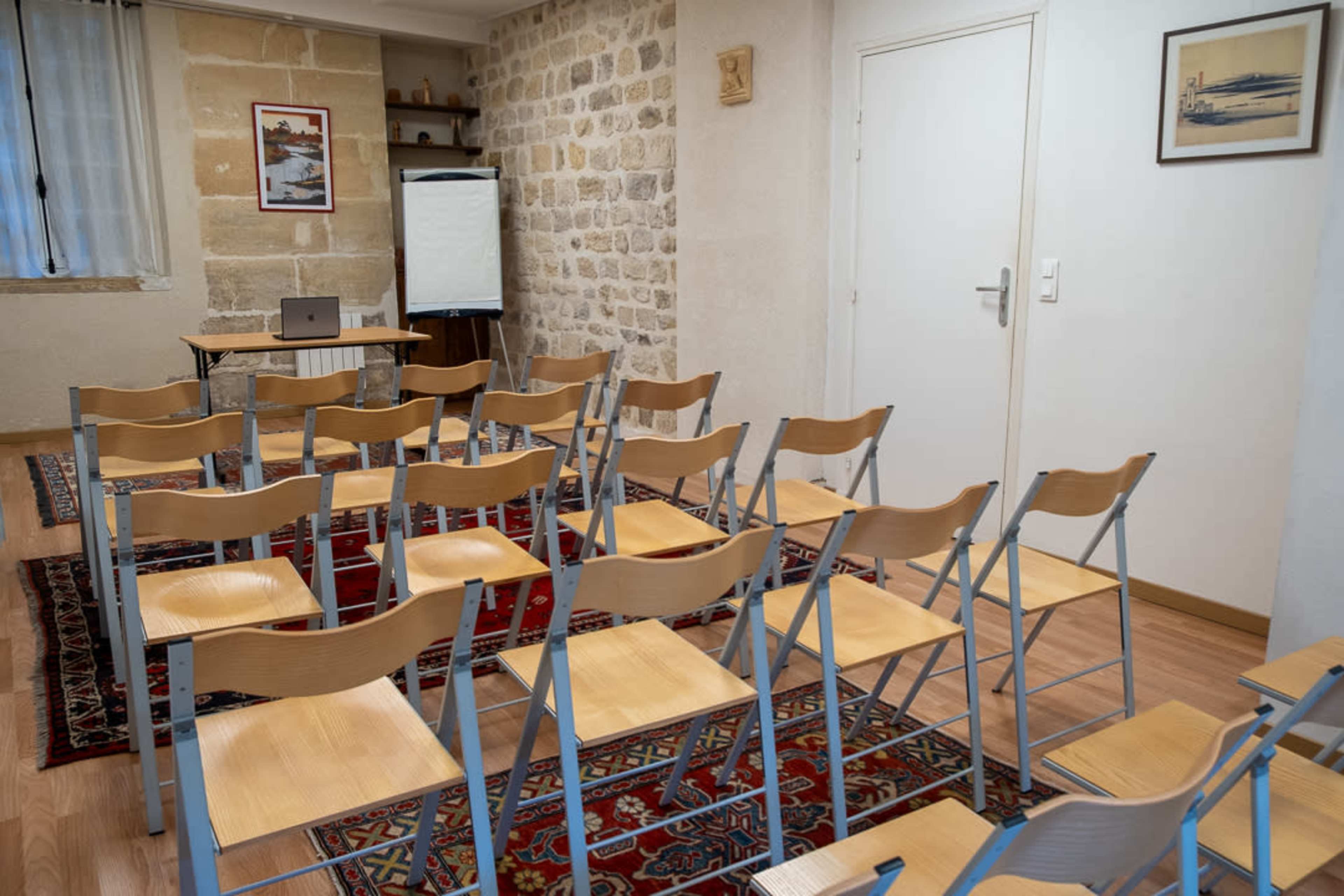 The image shows a classroom setup with rows of wooden chairs arranged in front of a table and a whiteboard in a room with stone walls.