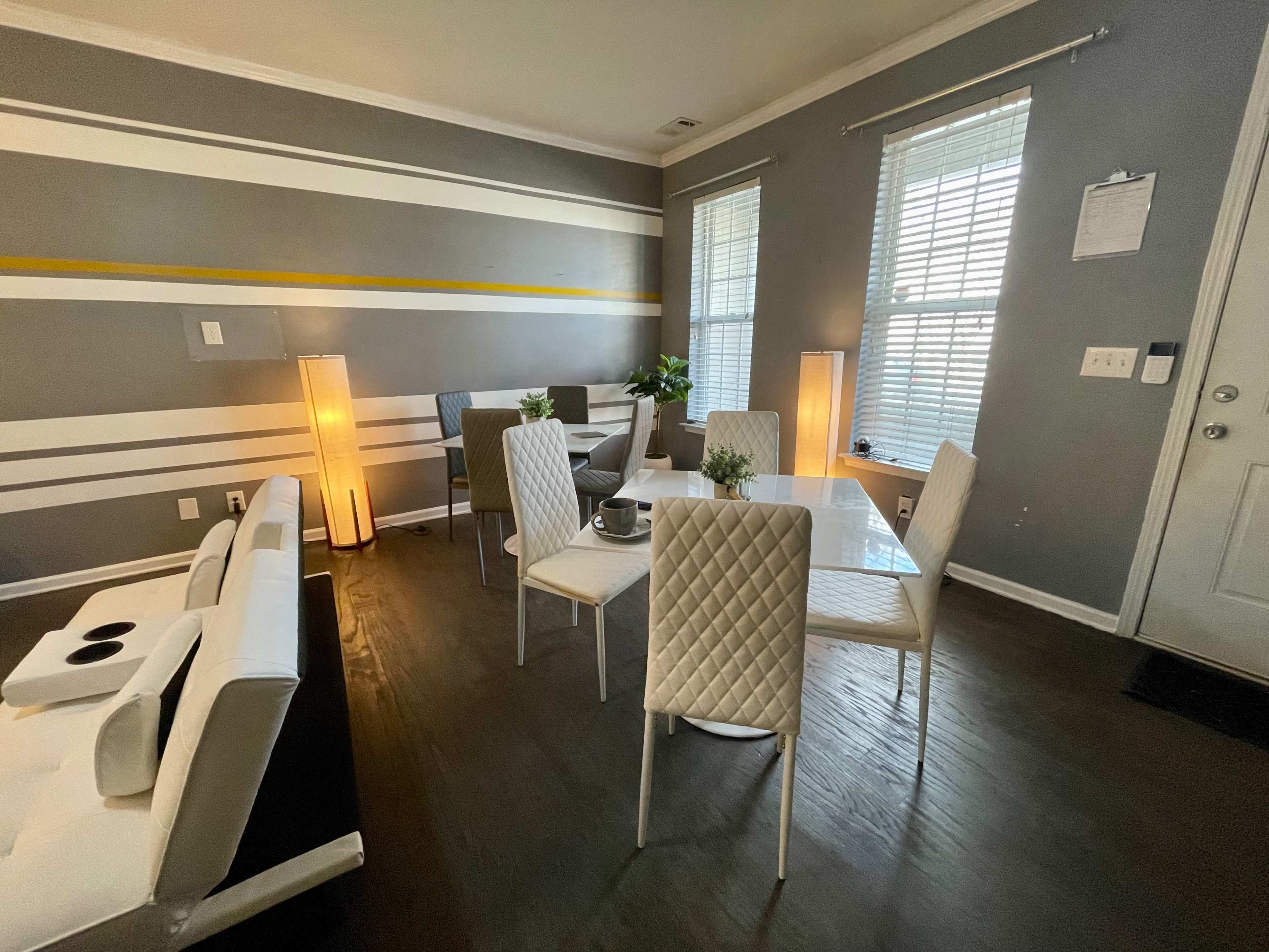 The image shows a modern dining area with a glass table, white chairs, and a sofa, featuring gray walls with horizontal stripes and two floor lamps.