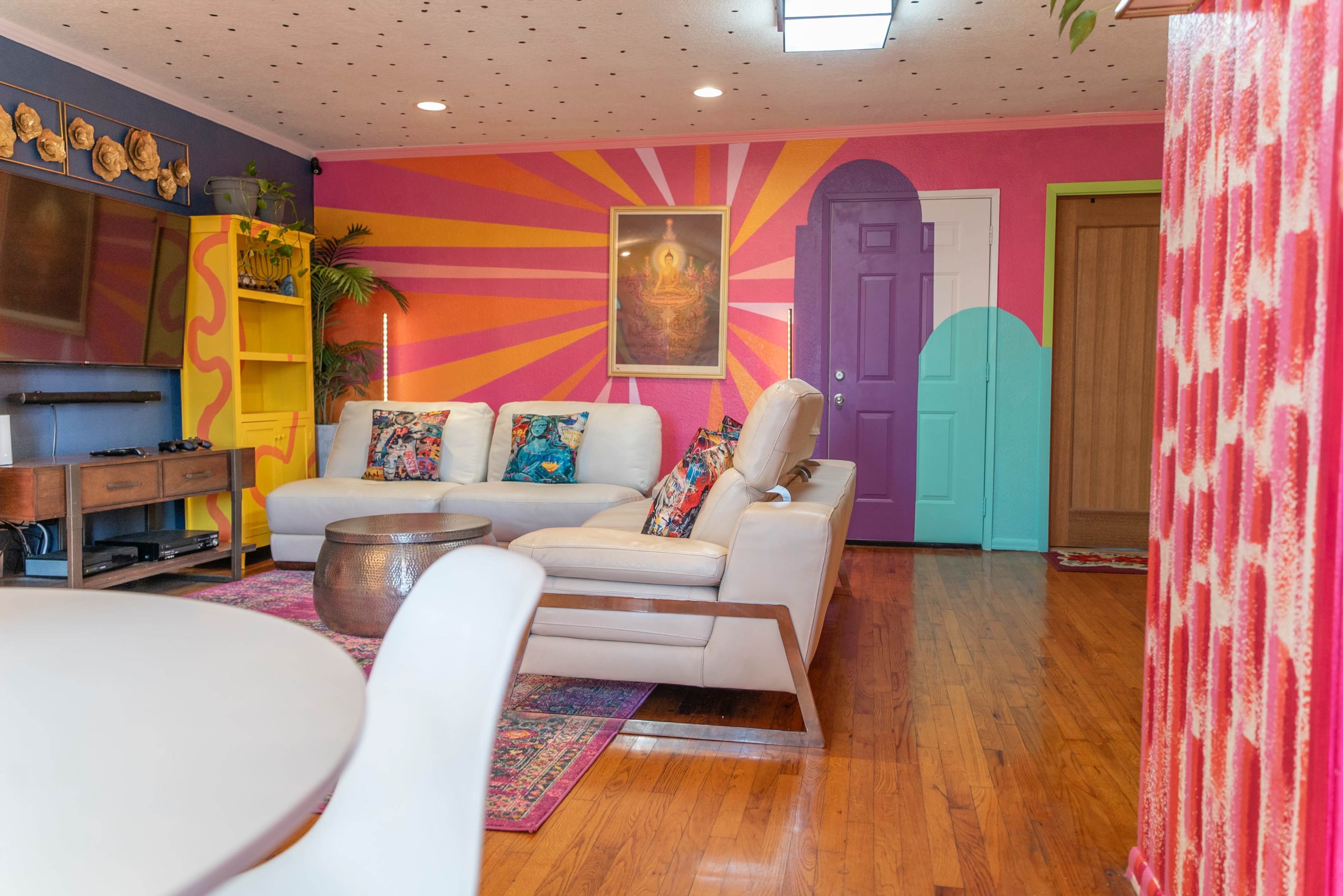 The image shows a brightly colored living room with a sectional sofa, decorative pillows, and vibrant wall art.