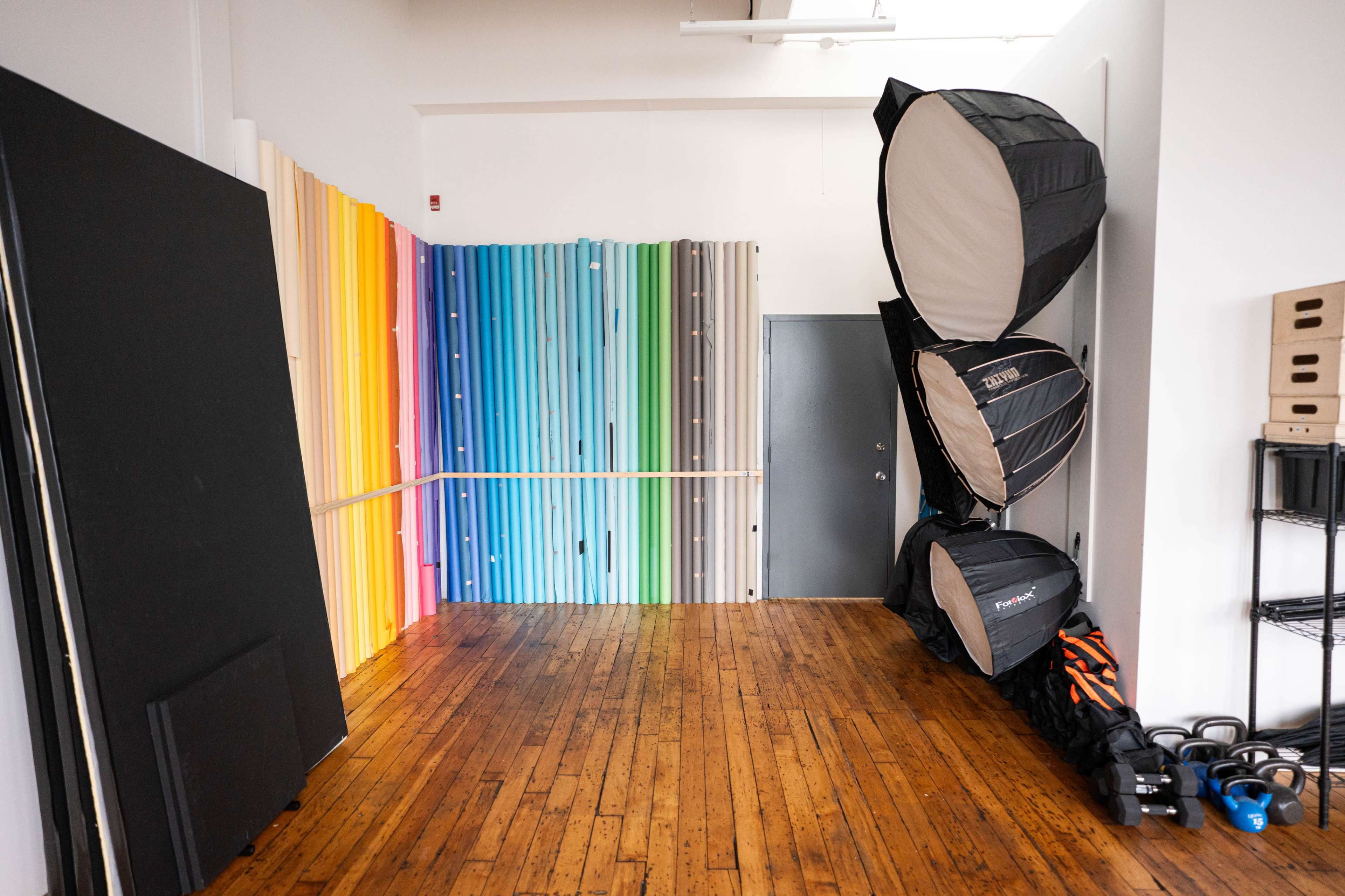 The image shows a bright studio space with a colorful backdrop composed of vertical rainbow-colored panels, a door on one side, and photography equipment lined up against the wall.