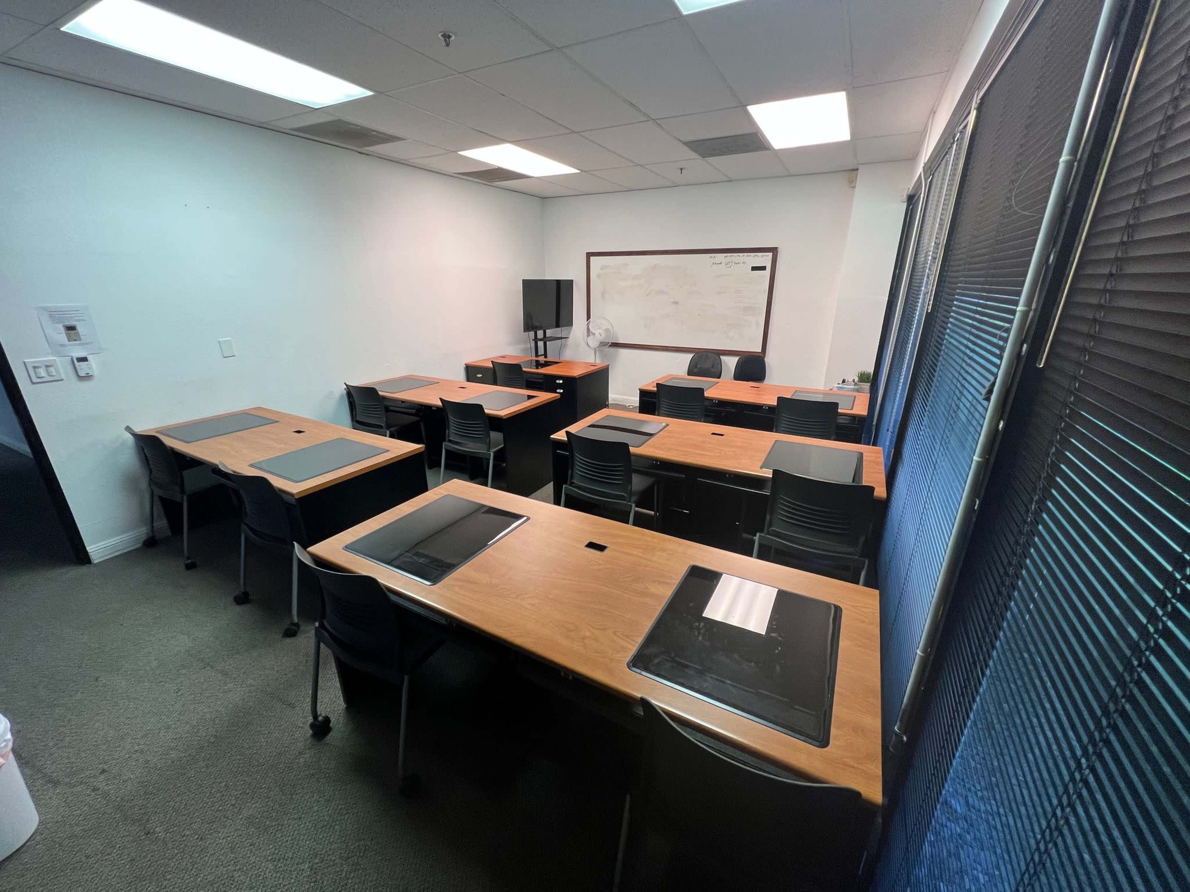 The image shows a sparsely furnished classroom with several wooden tables and black chairs arranged in rows, a whiteboard on the wall, and a television in the corner.