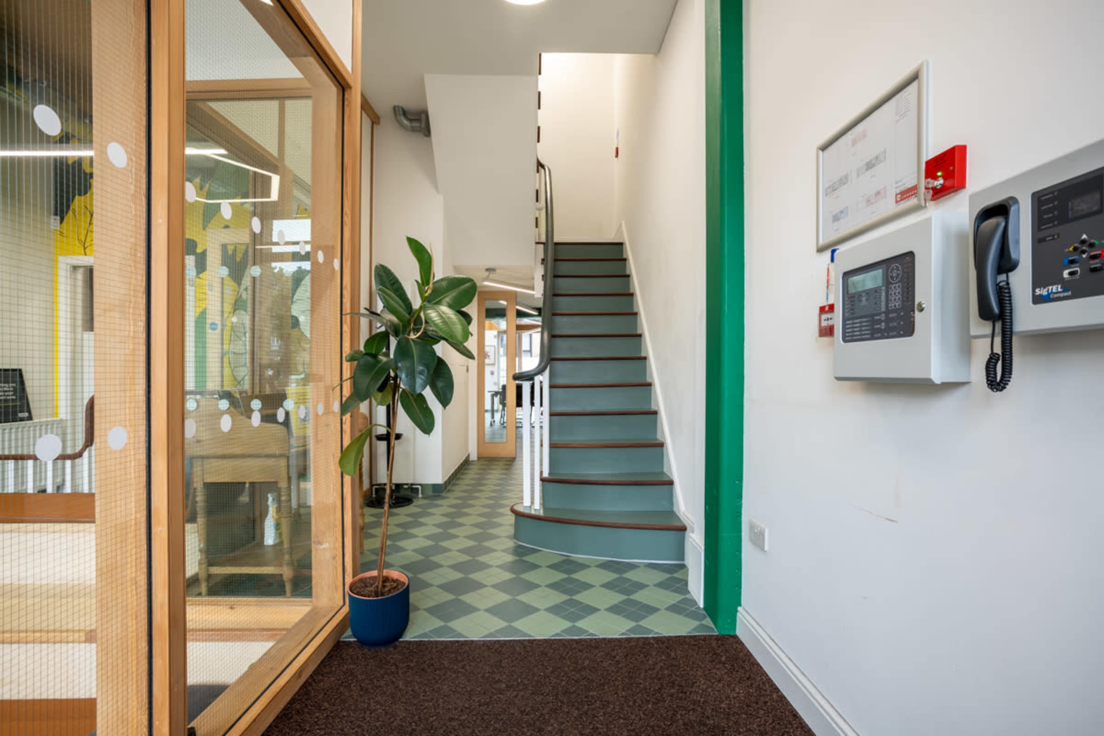 An interior hallway leading to a stairway, with a potted plant near the entrance and a wall-mounted intercom system.