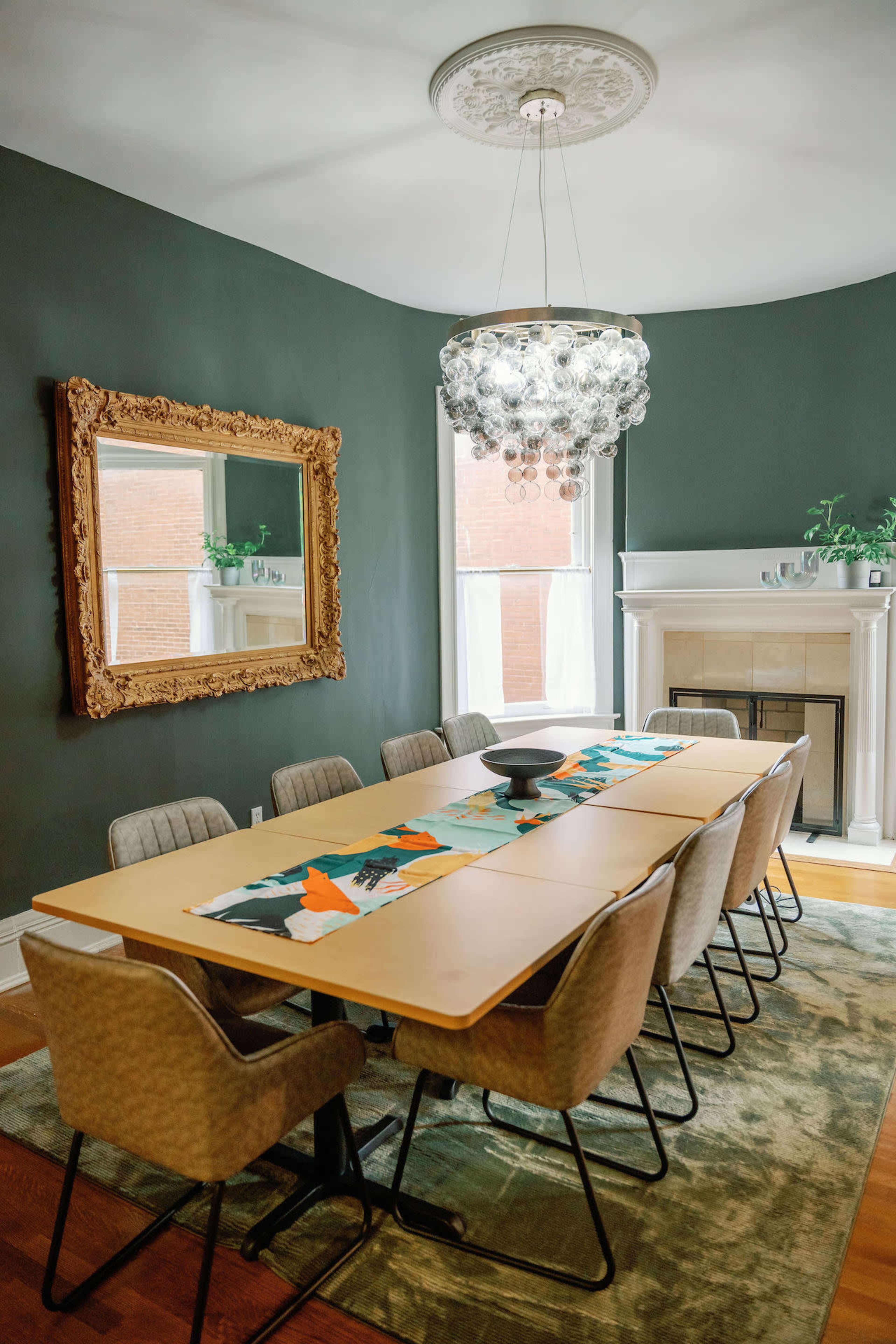 A large dining table is set in a stylish room featuring a decorative mirror, an elegant chandelier, and a fireplace.