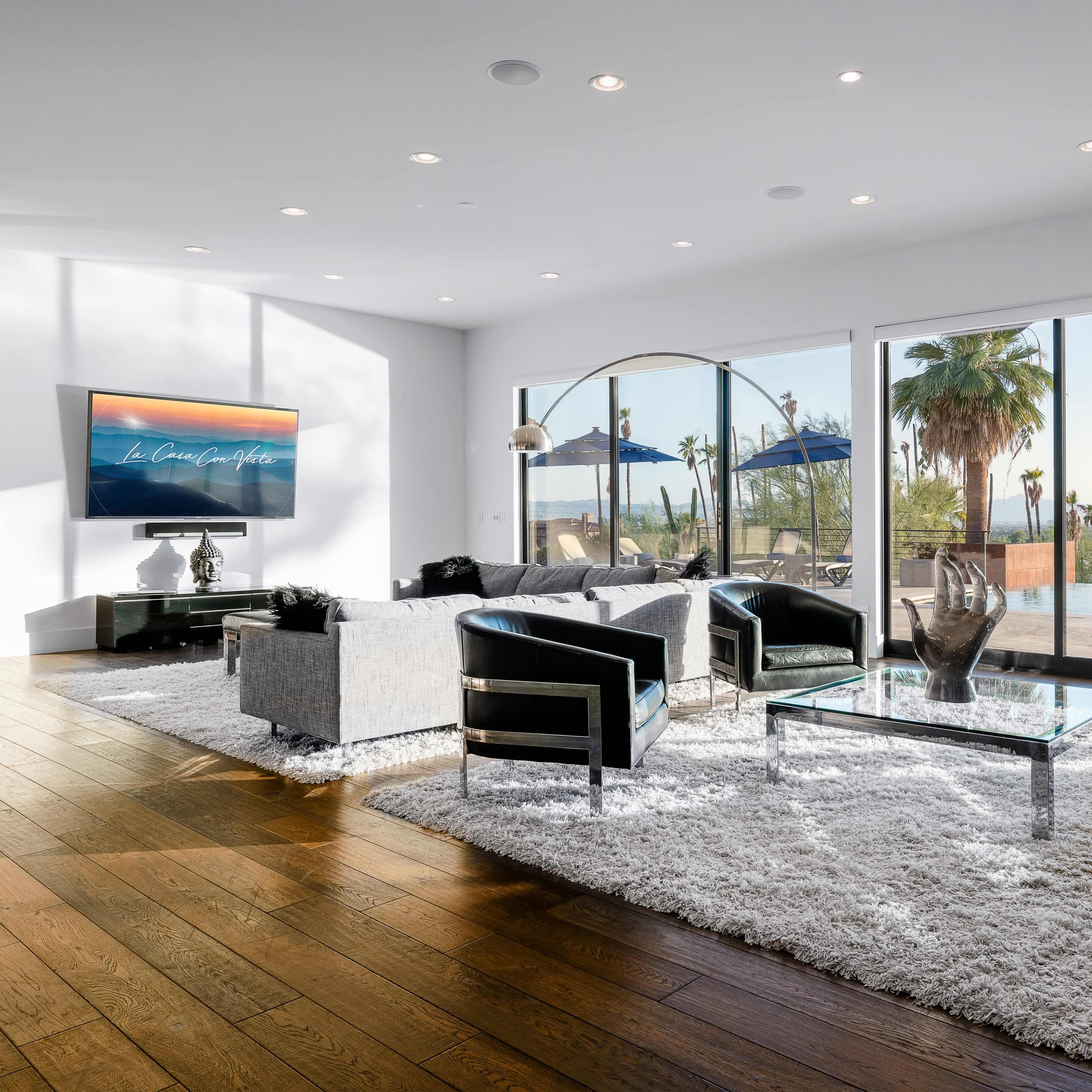 A modern living room with black and silver furniture, a television mounted on the wall, and large windows offering a view of palm trees and outdoor seating.