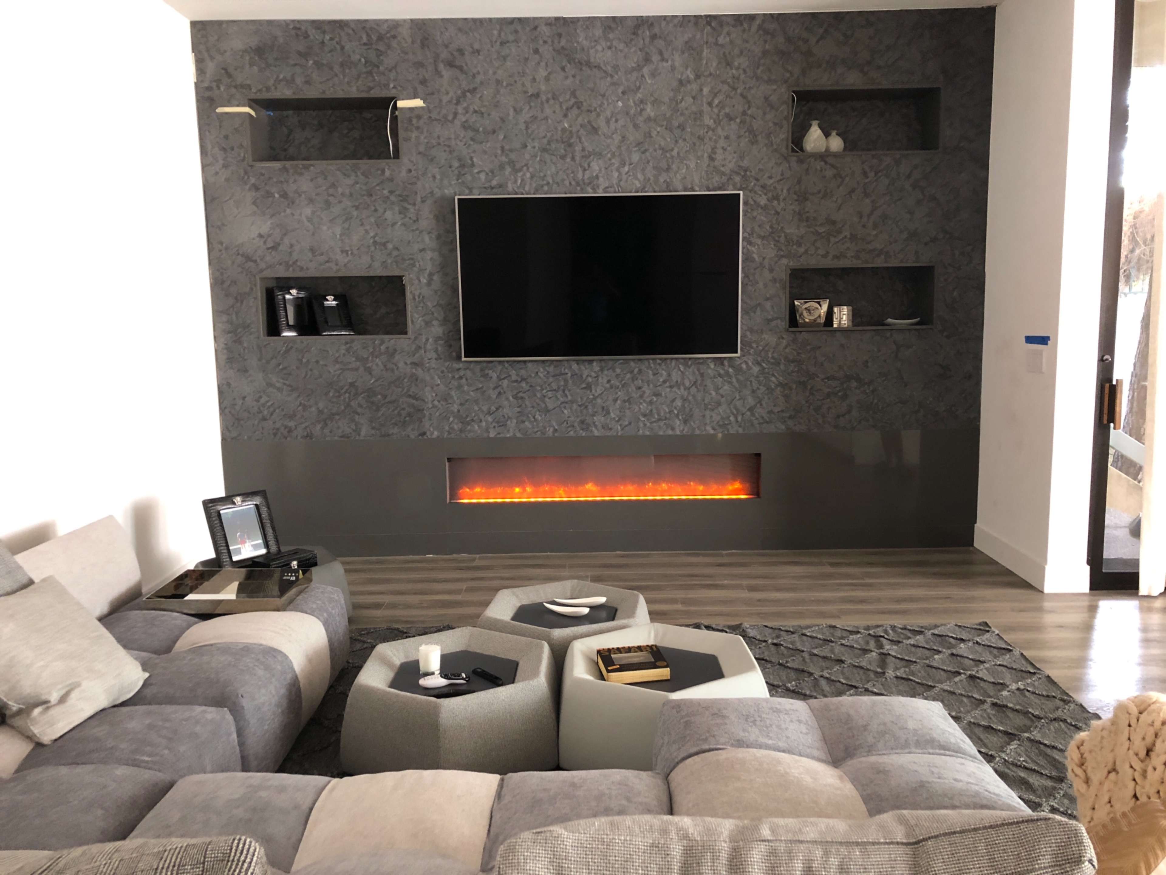 The image shows a modern living room with a large television mounted on a textured wall, flanked by minimalist shelves and an electric fireplace below, surrounded by a sectional sofa and geometric coffee tables.