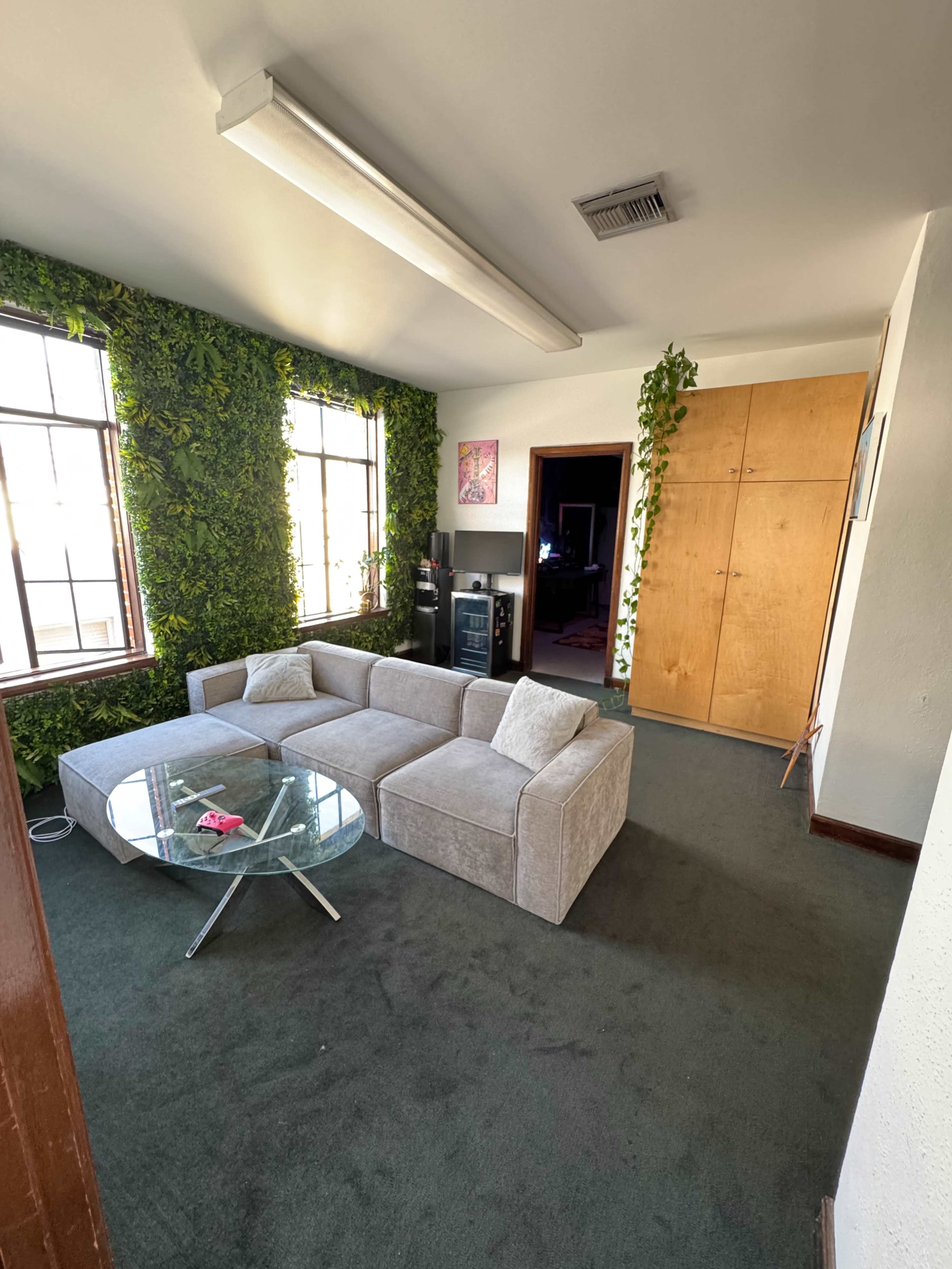 The image shows a modern living room with a beige sectional sofa, a glass coffee table, and greenery on the walls, featuring large windows and a door leading to another room.