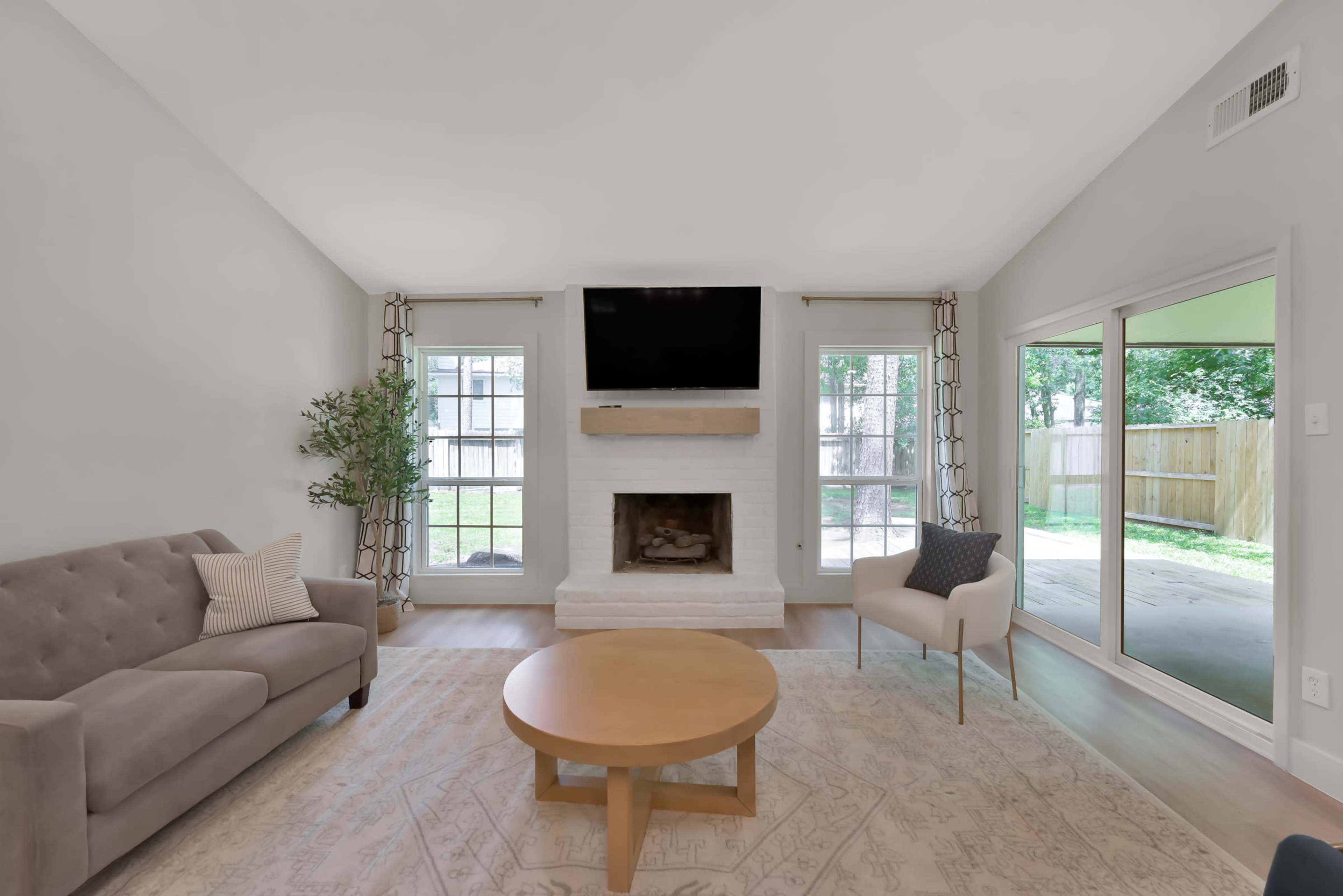 A living room with a gray sofa, a round wooden coffee table, a fireplace underneath a mounted TV, and large windows overlooking a backyard.
