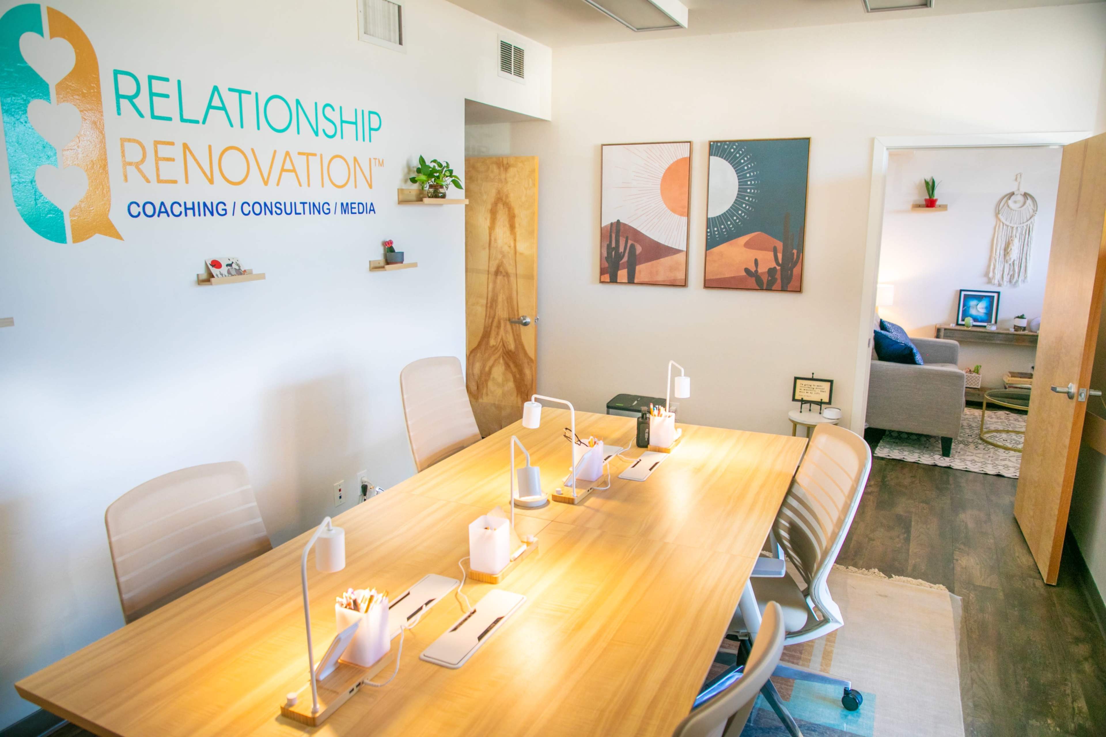 A modern meeting room with a large wooden table, several desk lamps, and walls adorned with artwork related to a coaching and consulting business.