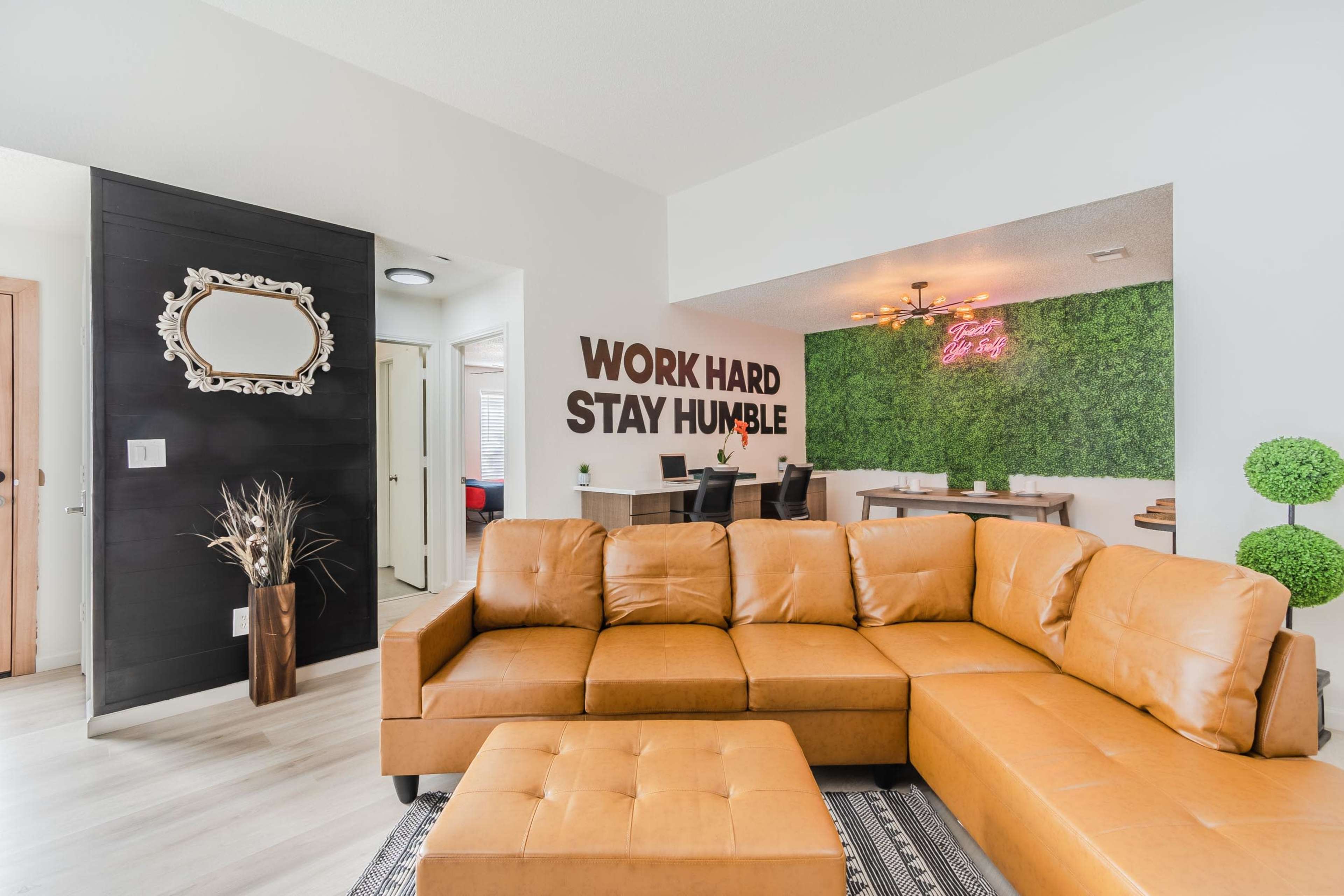 A modern living room features a brown leather sectional sofa facing a wall with the words "WORK HARD STAY HUMBLE" and greenery decor in the background.