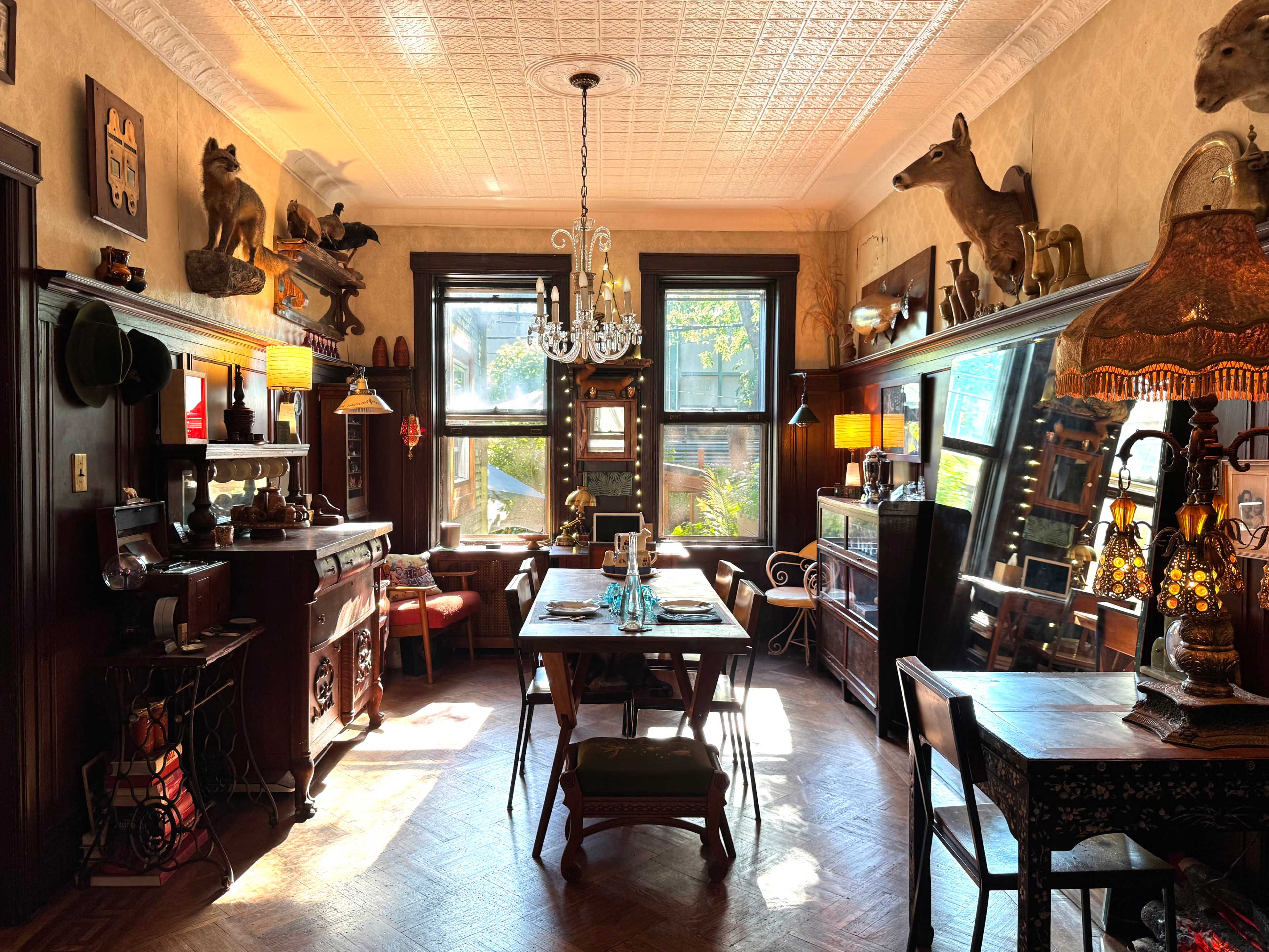 The image shows a vintage dining room filled with ornate furniture, animal mounts on the walls, and large windows allowing natural light to illuminate the space.