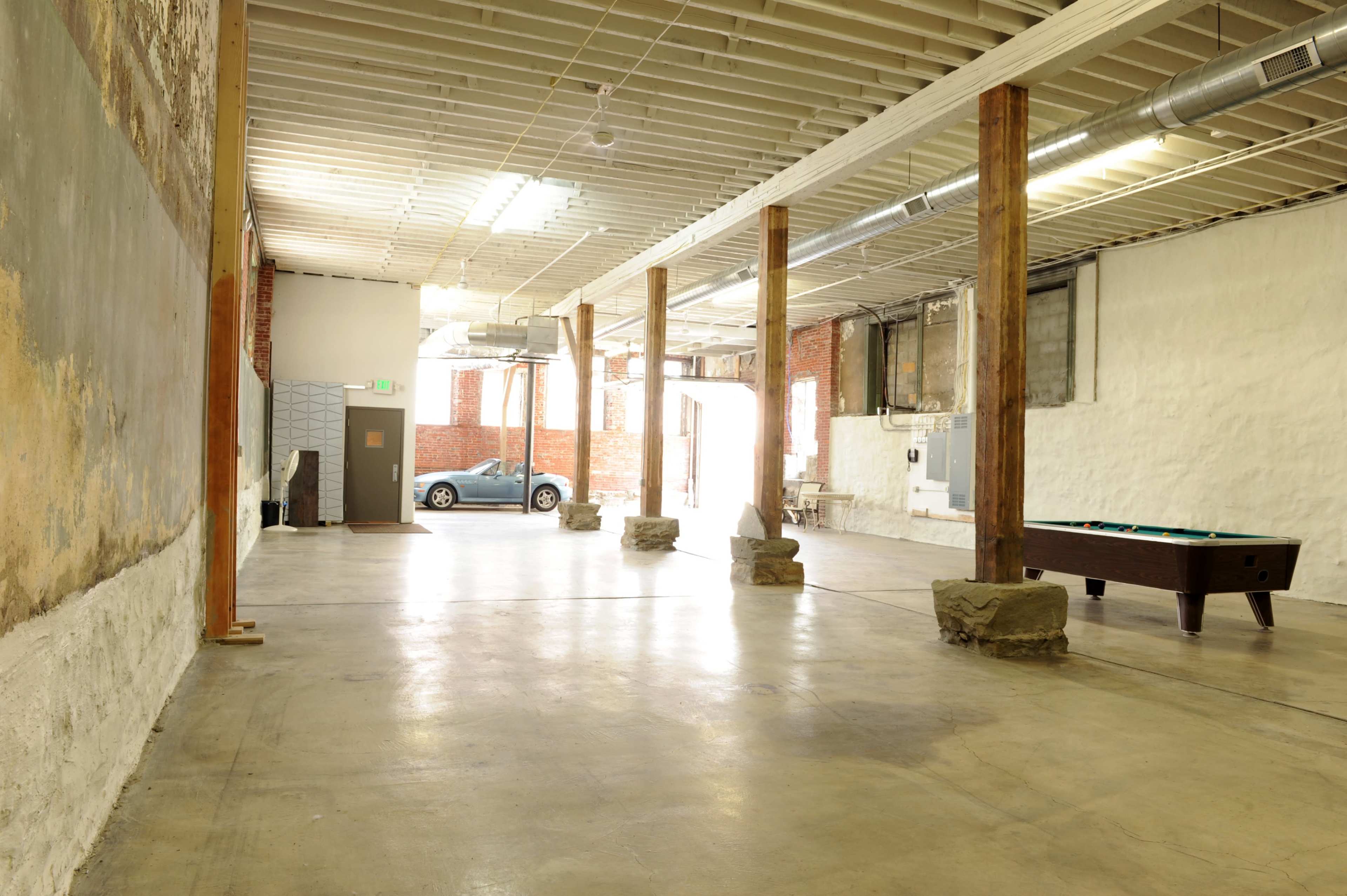 The image shows a spacious, vacant interior with exposed walls, wooden columns, and a pool table in the center.