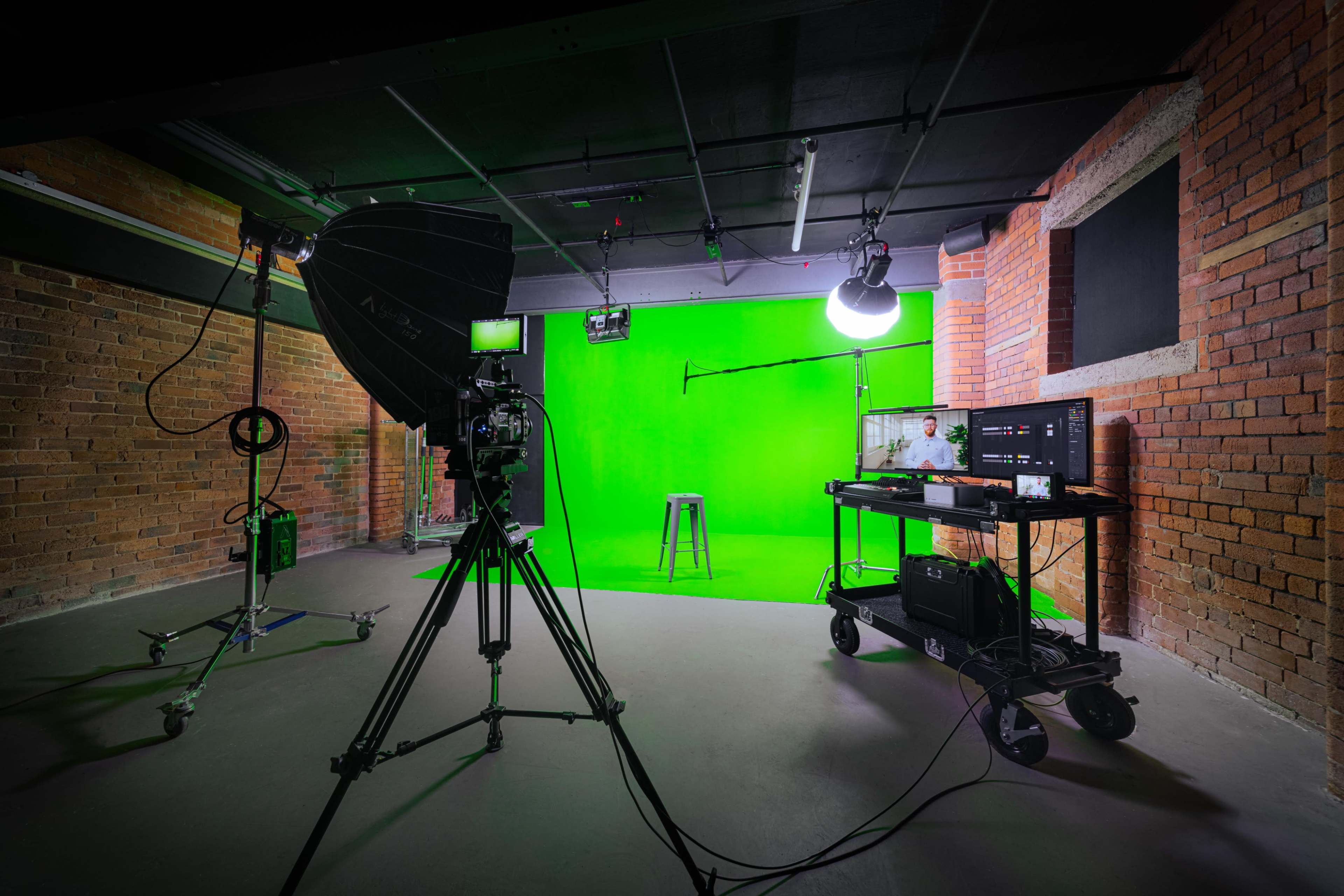 A studio setup featuring a green screen, camera equipment, lighting, and a stool in a brick-walled room.