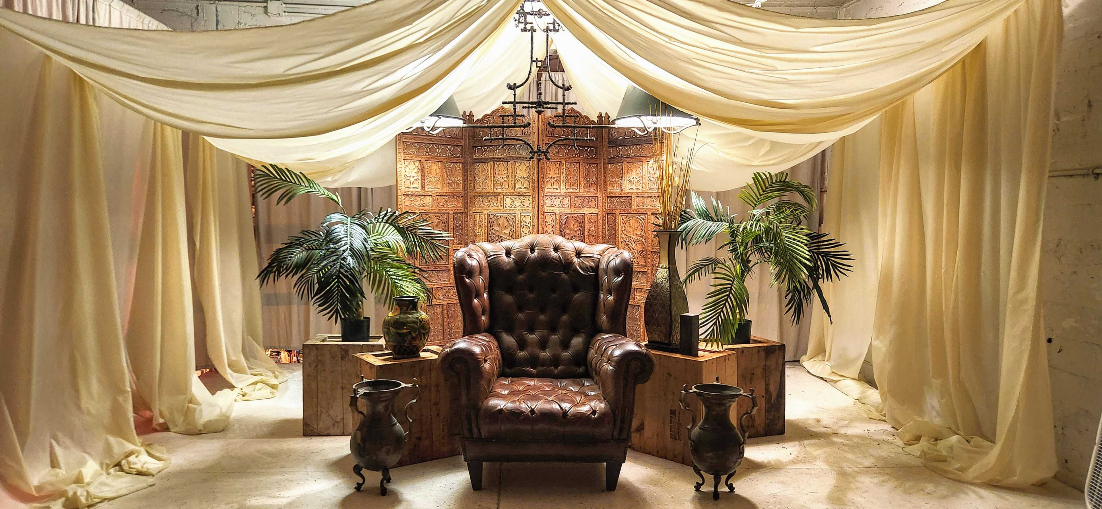 A brown leather armchair is surrounded by potted palm plants and wooden boxes beneath a draped canopy of fabric.