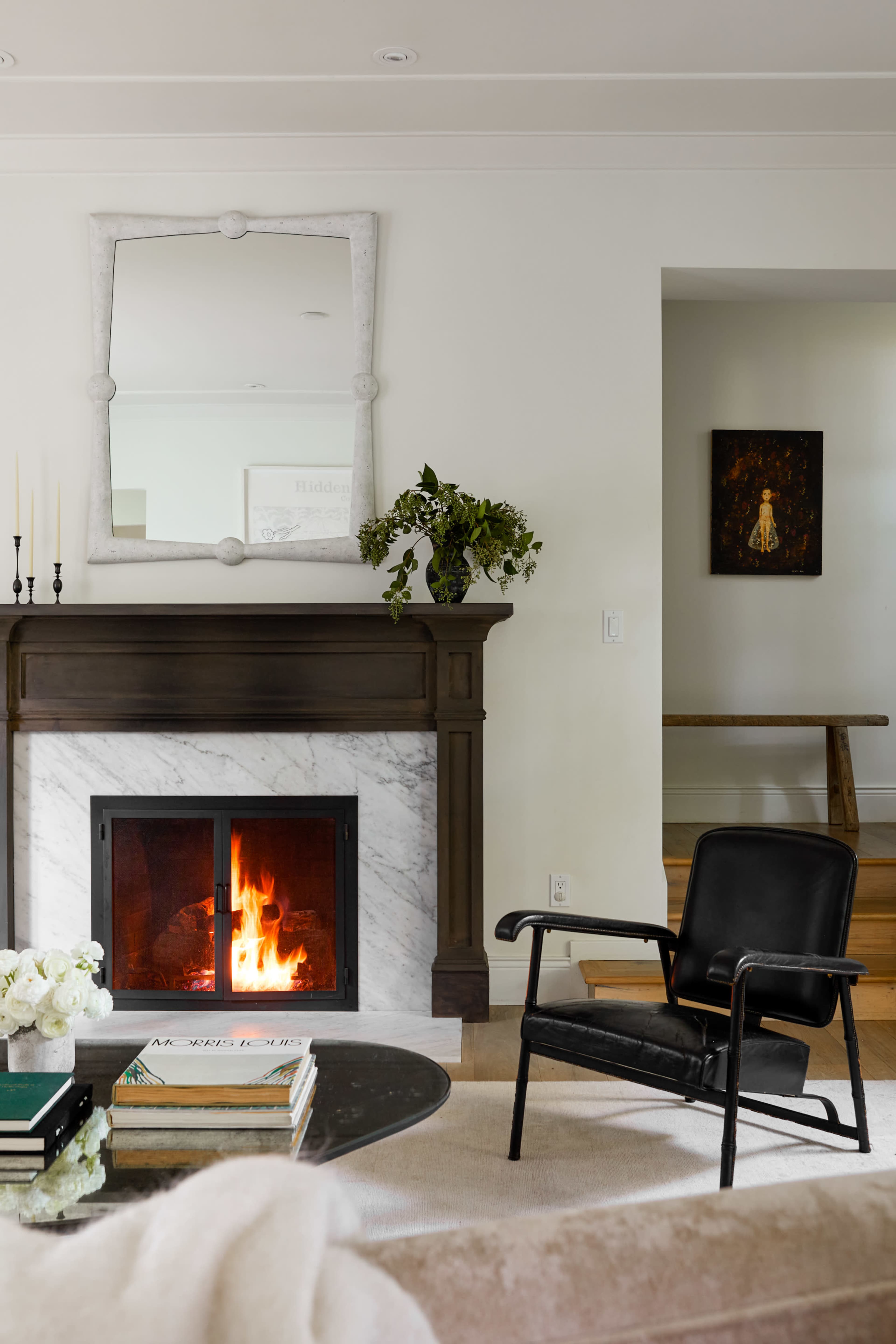 A black chair sits next to a marble fireplace, which has a fire burning and a mirror above it, while a small table with books and a vase of flowers is in the foreground.