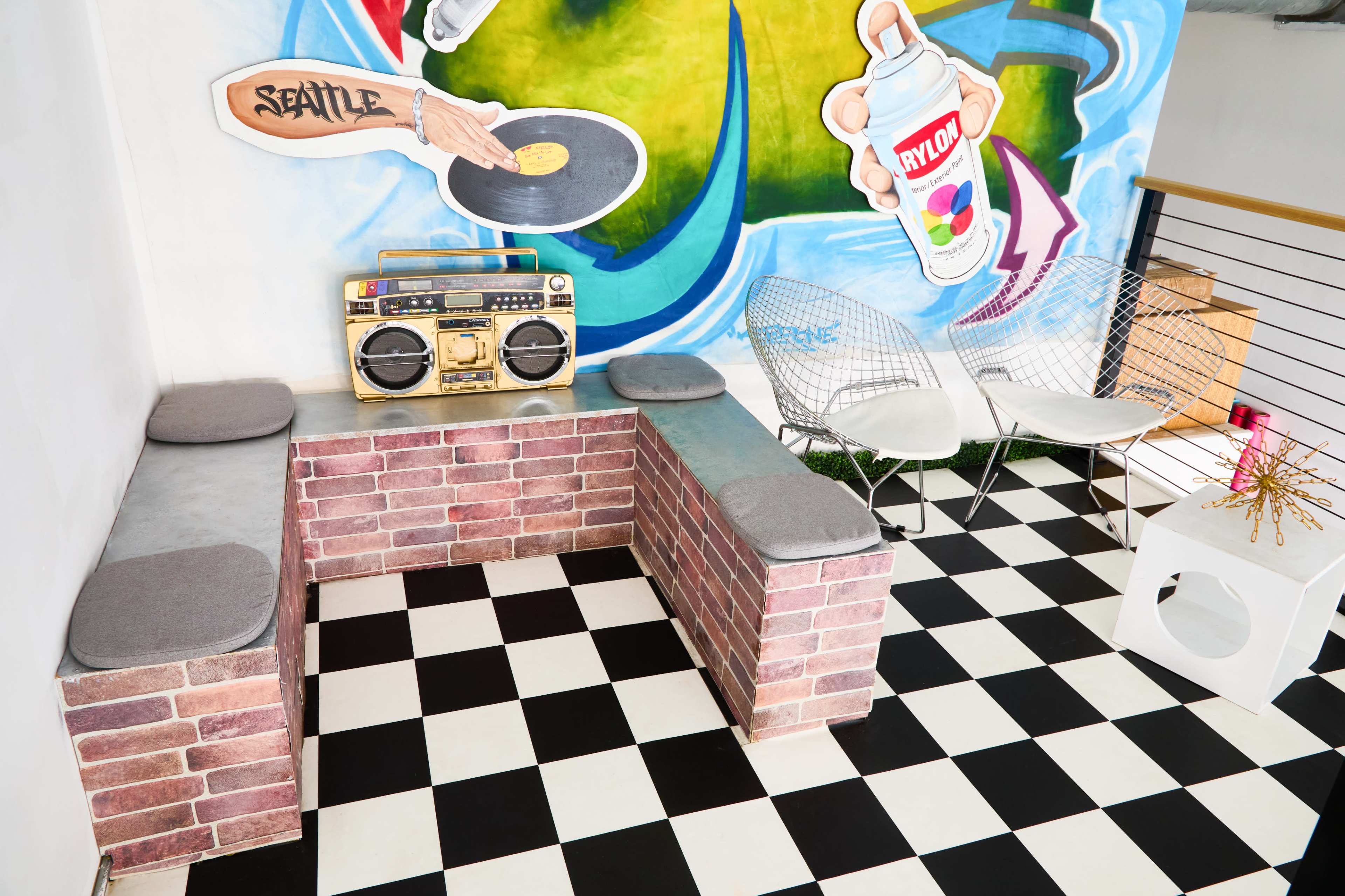 The image shows a vibrant lounge area featuring a brick-patterned seating space, a retro boombox, and a colorful mural on the wall, with checkered flooring.