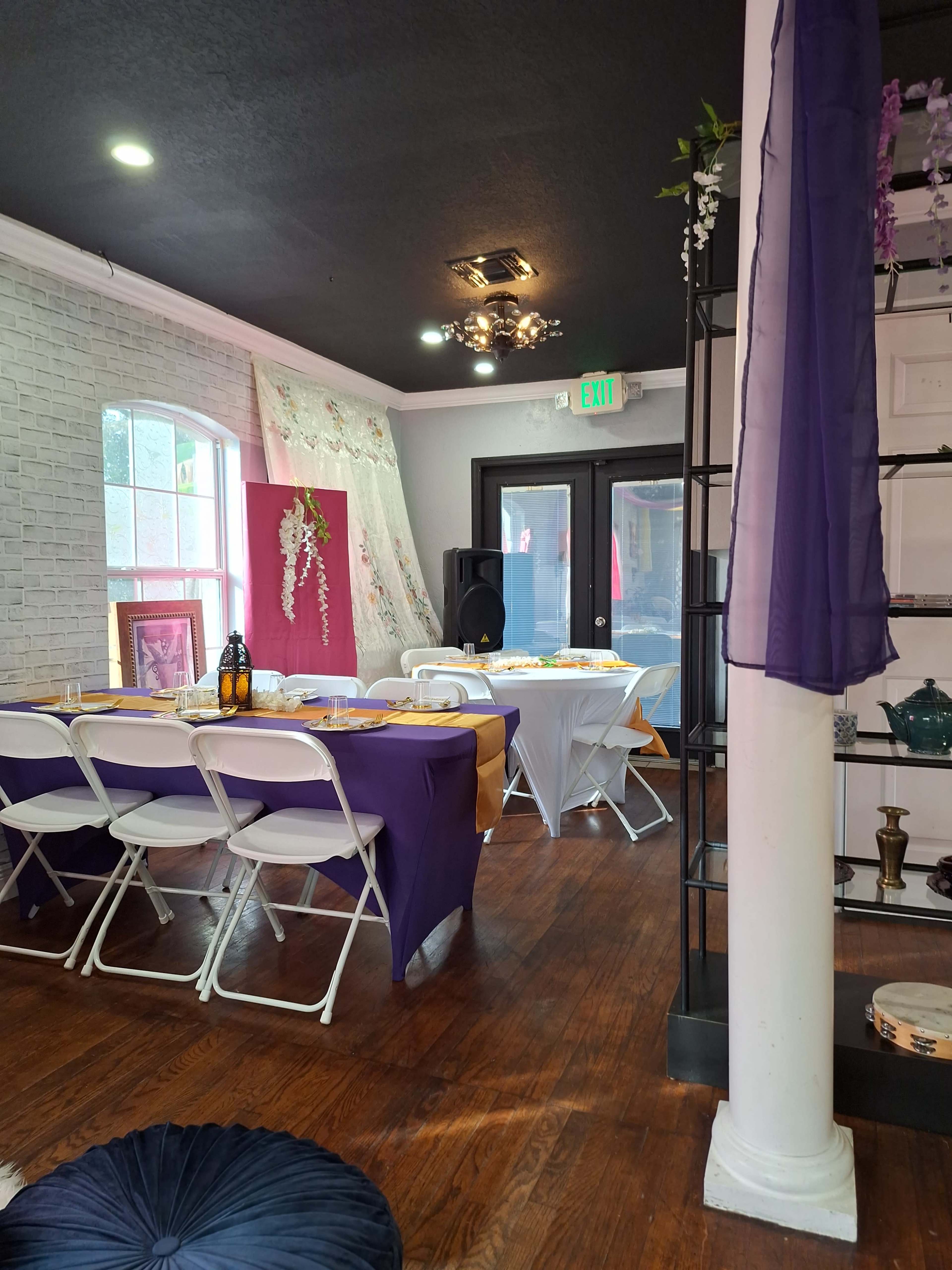 An elegantly decorated event space featuring tables set with purple and white tablecloths, a speaker system, and decorative elements against a brick wall.