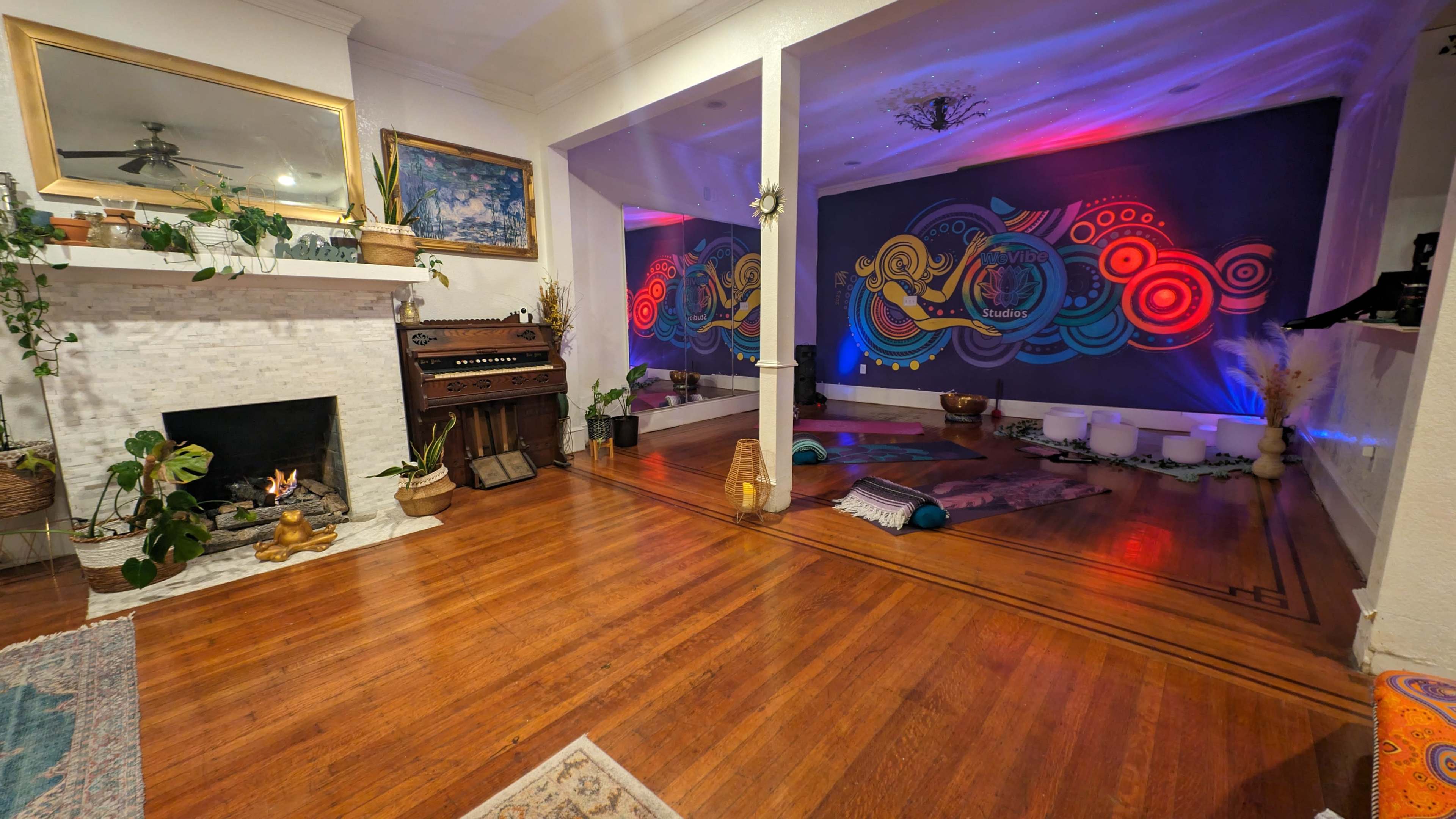 The room features a decorative fireplace and an adjacent area adorned with colorful murals, plants, and various seating arrangements on wooden flooring.