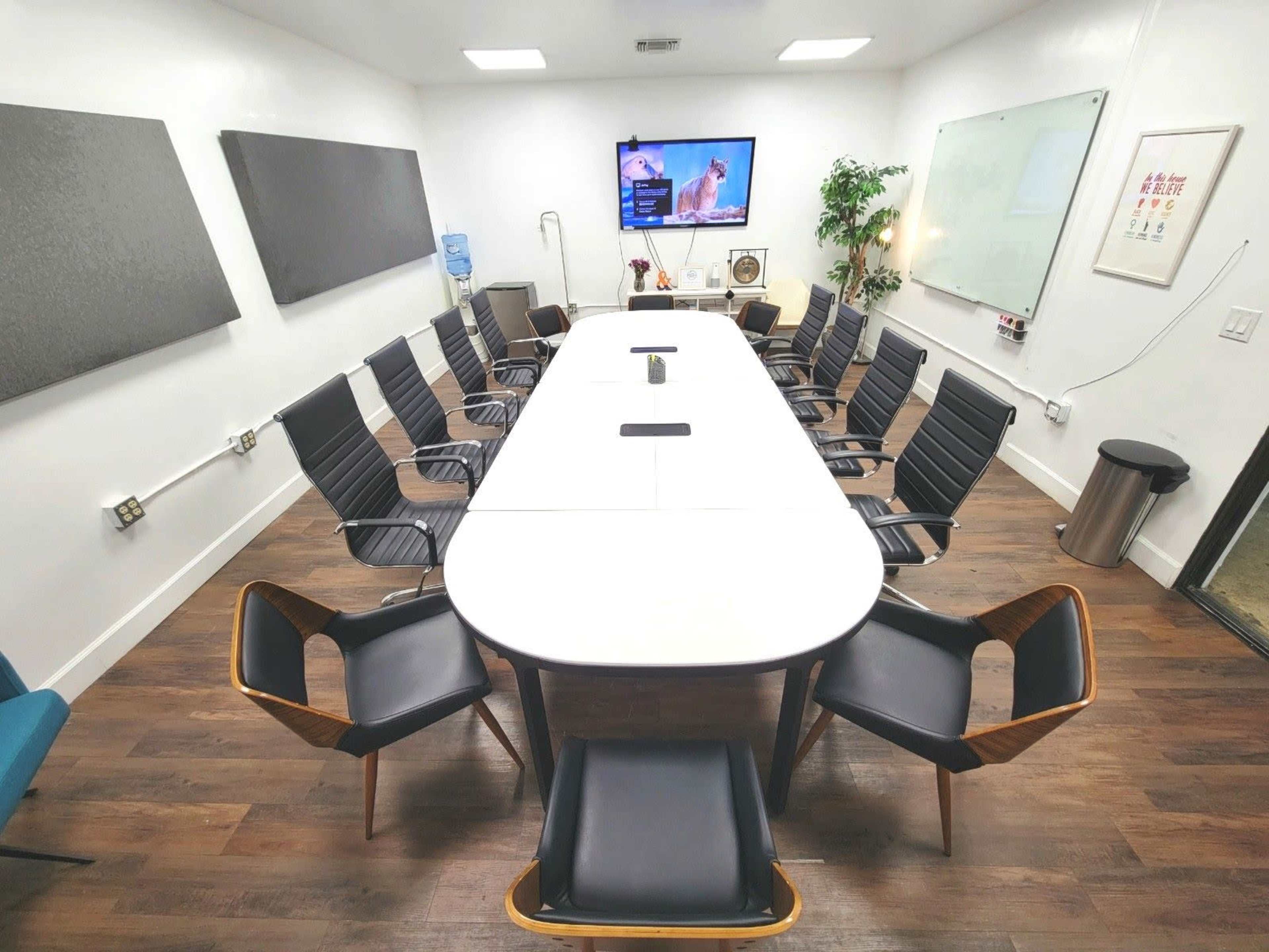 The image shows a modern conference room with a long white table surrounded by black and wooden chairs, a television mounted on the wall, and a whiteboard.