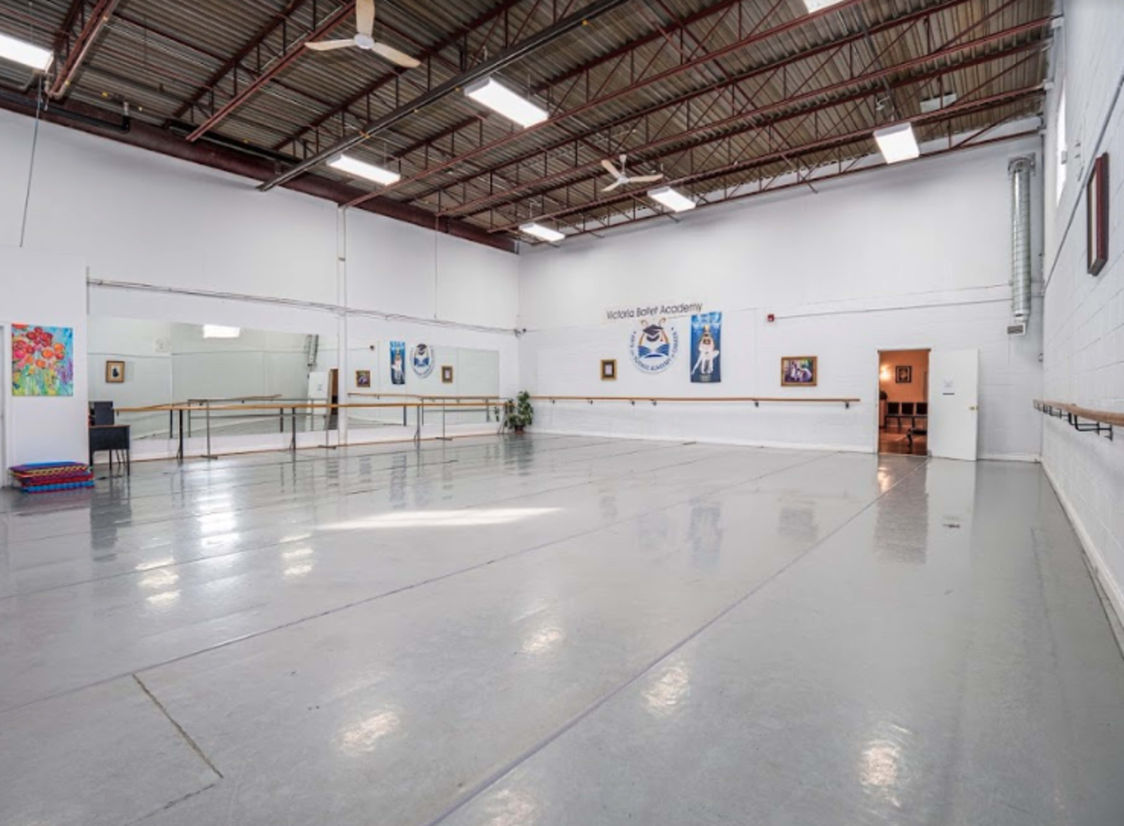An empty dance studio features a mirrored wall, ballet barres, and a concrete floor, with artwork displayed on the walls.