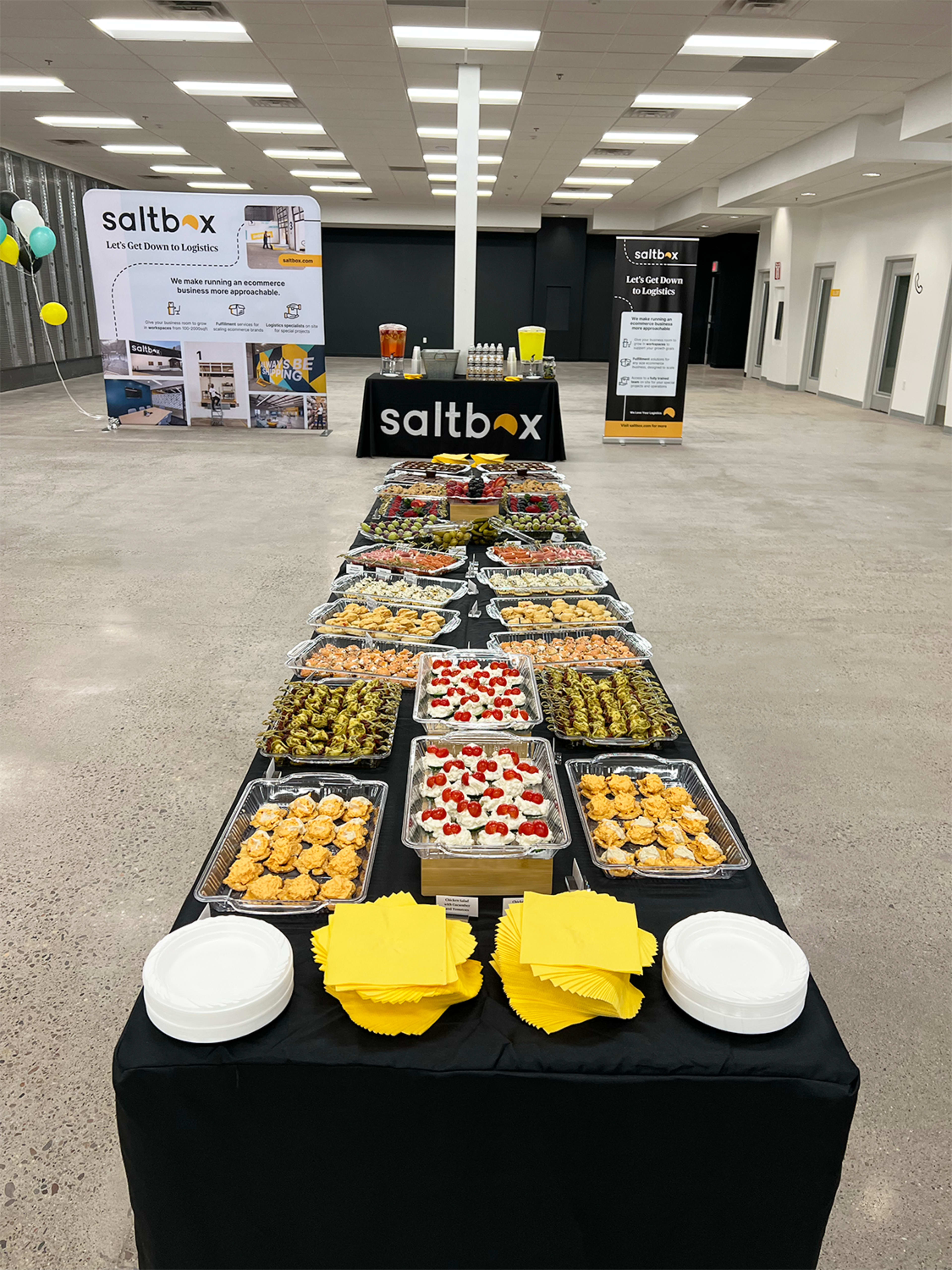A long table is filled with a variety of food platters, including desserts and appetizers, under a banner for Saltbox in a spacious indoor setting.