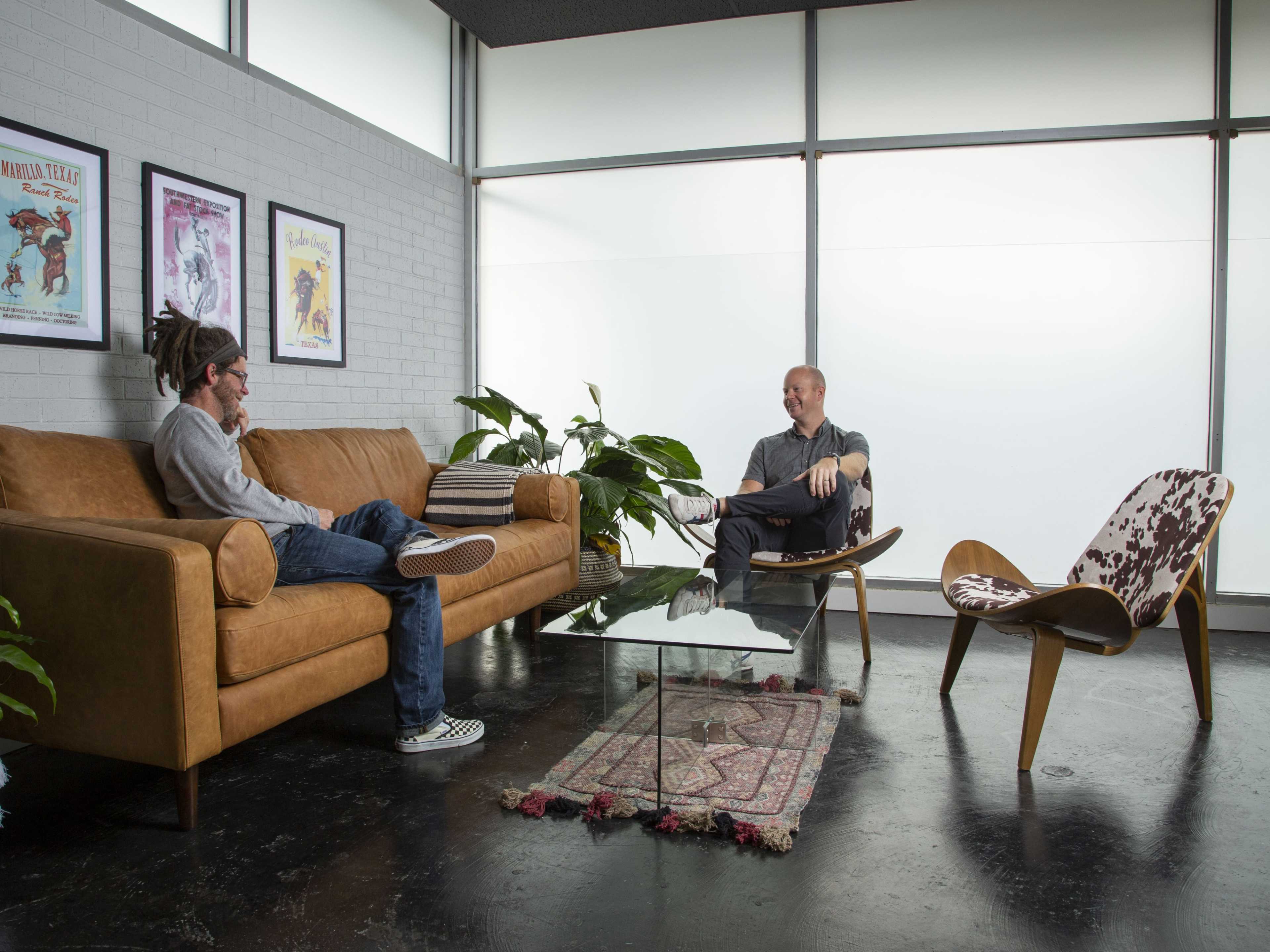 Two men are seated in a modern room, one on a brown leather sofa and the other in a chair, with plants and a glass coffee table between them.