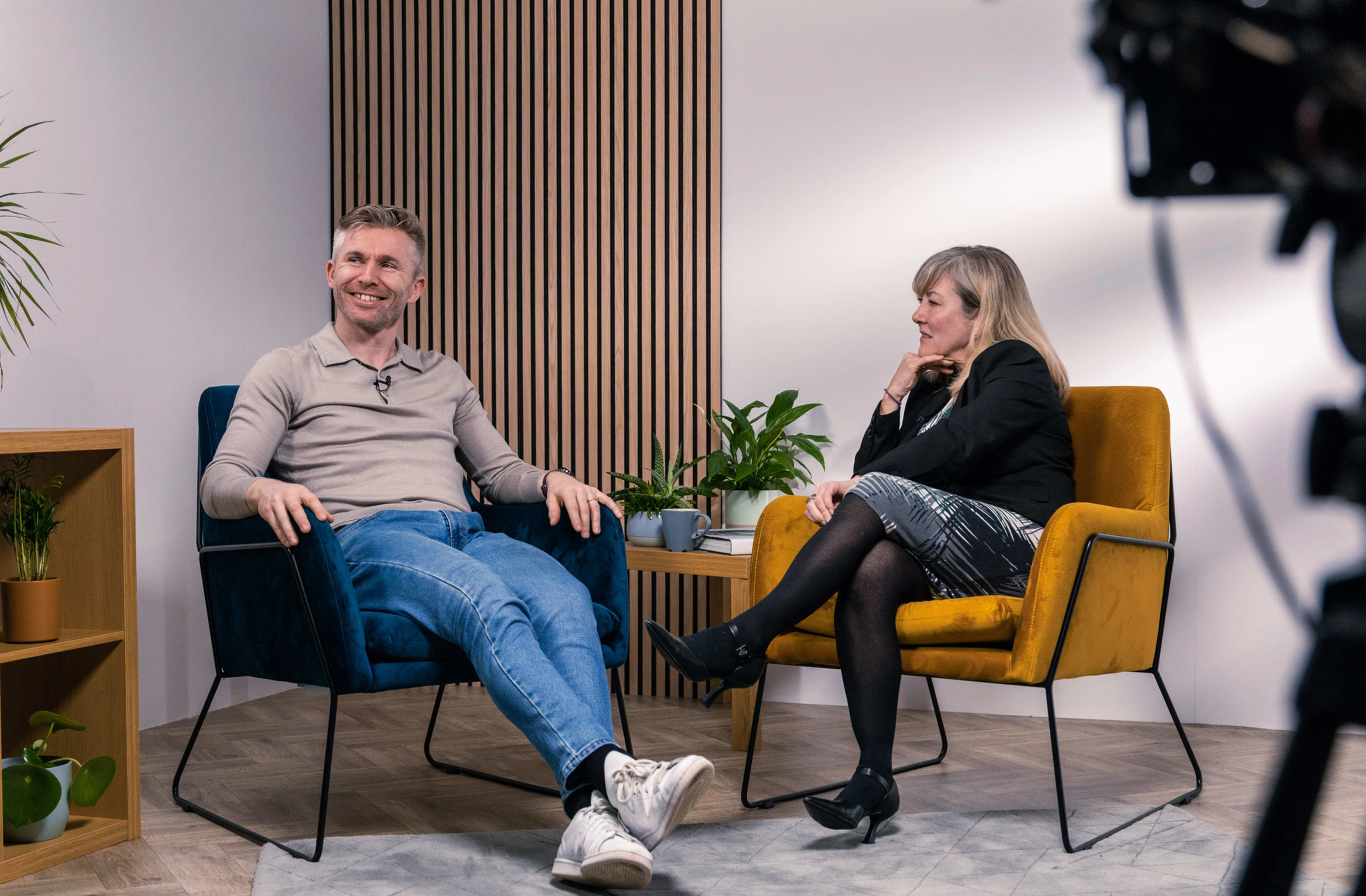 A man and a woman are seated in modern chairs during a discussion in a well-lit studio with plants and wooden accents in the background.