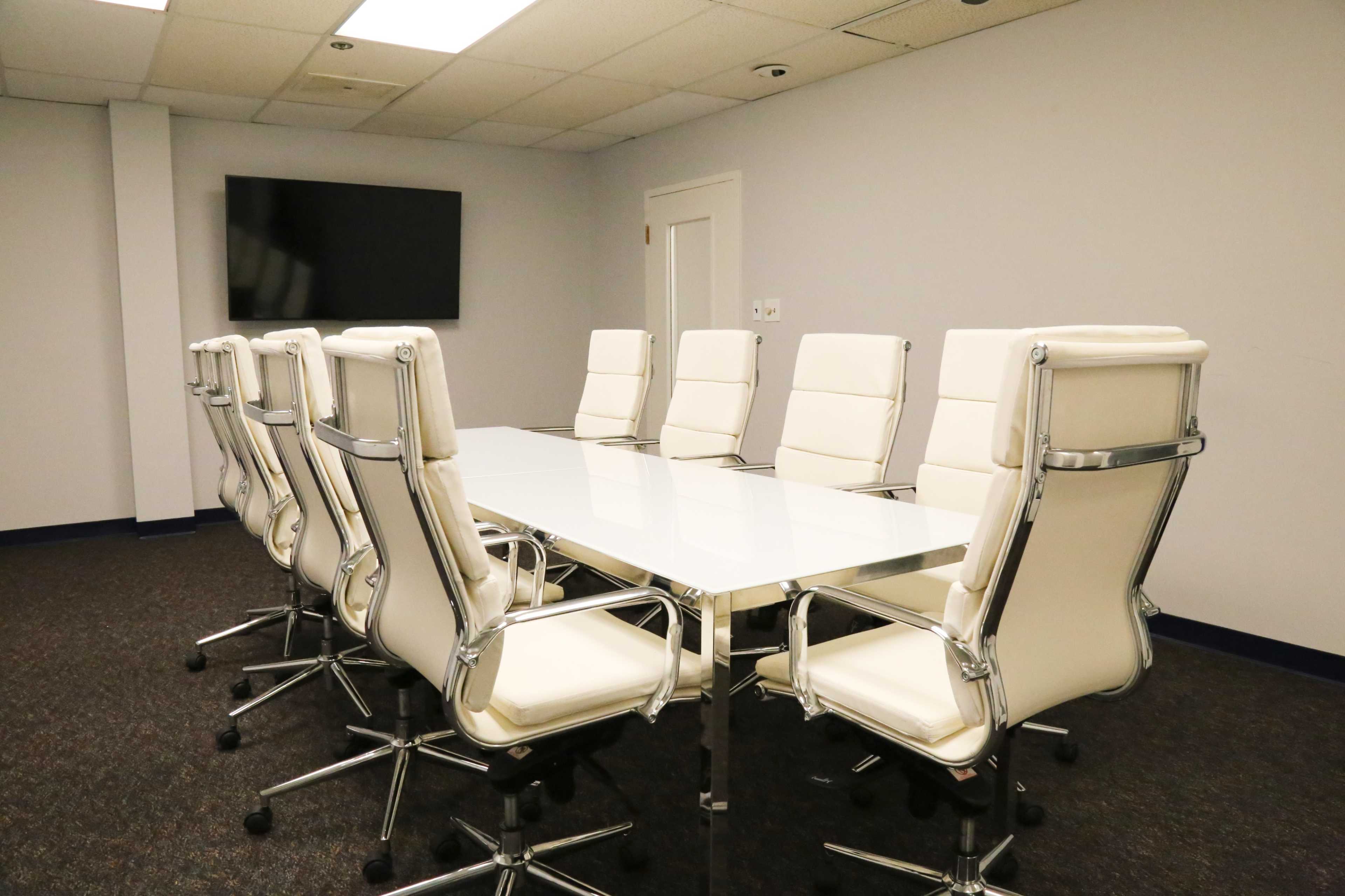A modern conference room features a long glass table with white leather chairs arranged around it and a television mounted on the wall.