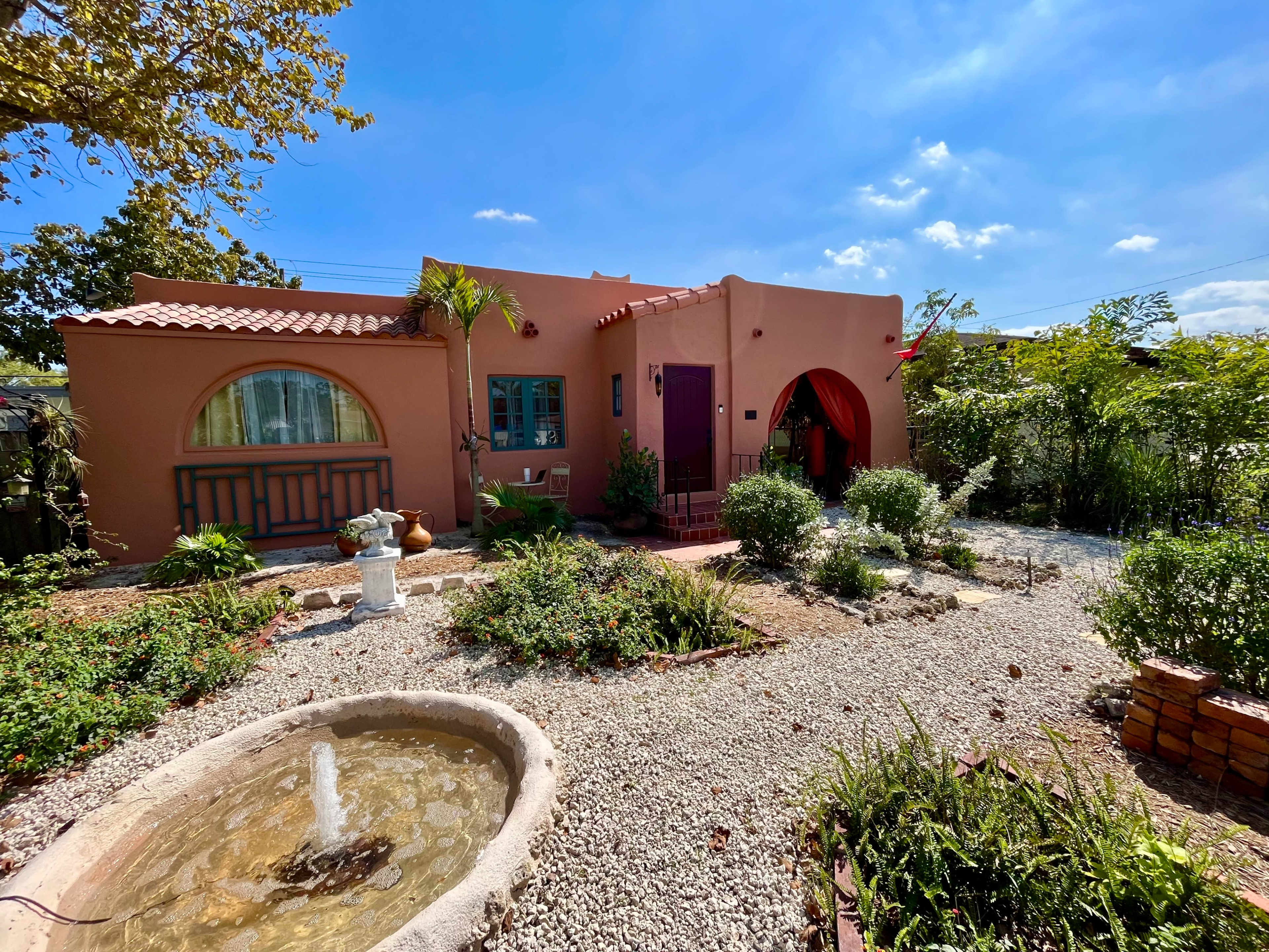 A pink stucco house with arched windows and a red door is surrounded by a garden featuring a stone fountain and various plants.
