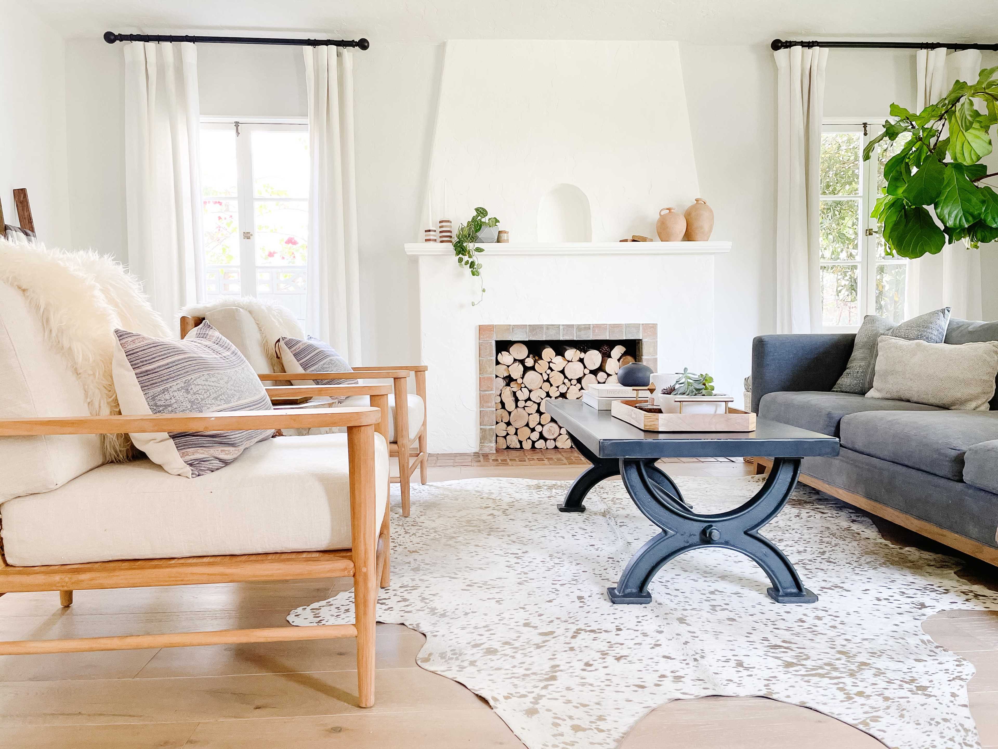 A cozy living room features a light-colored rug, a wooden coffee table, two armchairs, a gray sofa, and a fireplace with stacked logs, enhanced by natural light from the windows.