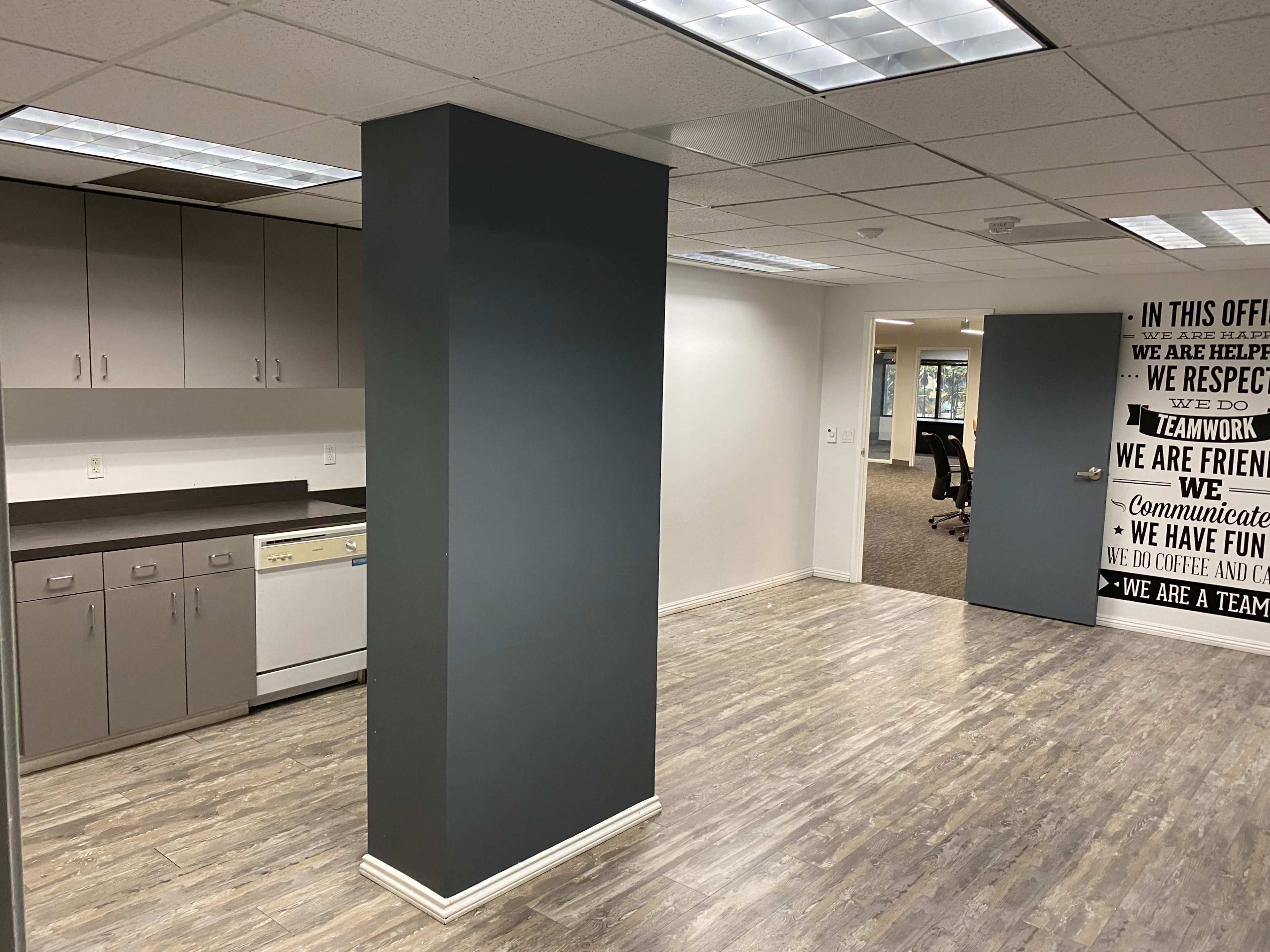 An office space featuring a kitchenette area with cabinets and a dishwasher, a central column, and a wall adorned with motivational text.